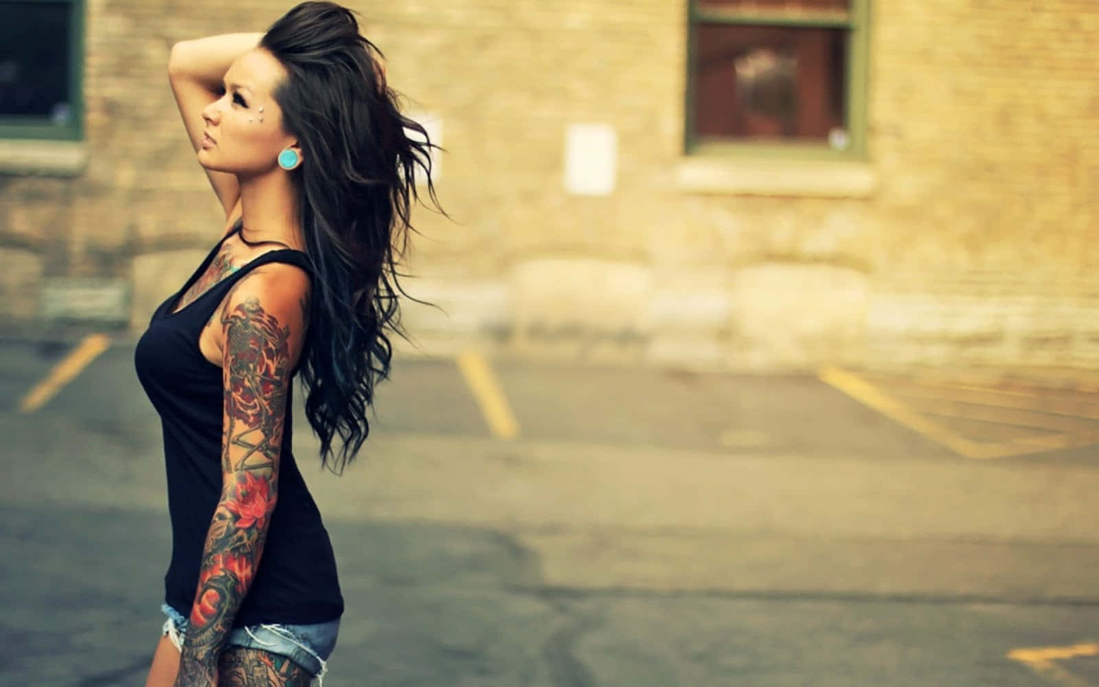 Tattoo Girl 桌布 壁紙 — 一位刺青黑髮女性站在陽光普照的城市小巷中，手臂舉起，風吹起她的髮絲。