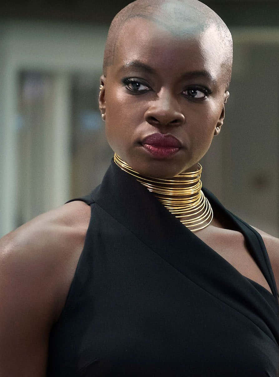 Dora Milaje 桌布 壁紙 — 一位剃光頭的優雅女士，戴著大膽的金色頸鏈和黑色連衣裙，身處柔和模糊的室內環境。
