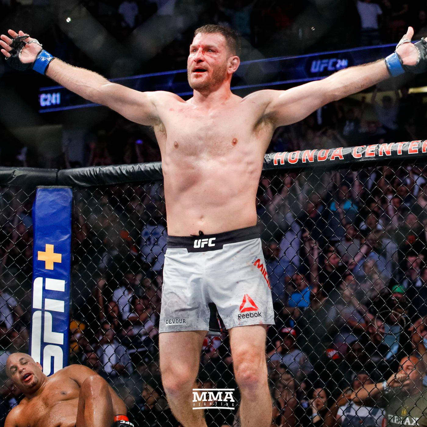 UFC 桌布 壁紙 — 一位勝利的MMA拳手在八角籠中高舉雙臂，慶祝勝利，而他的對手則在明亮的場館燈光下倒地敗北。