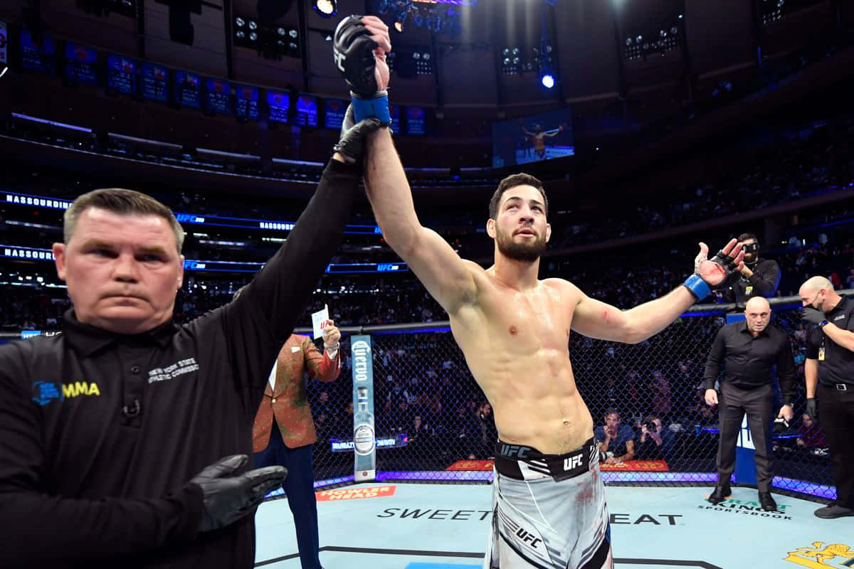 Victory 桌布 壁紙 — 一位勝利的MMA拳手在燈光明亮的八角籠內慶祝，裁判舉起他的手臂。