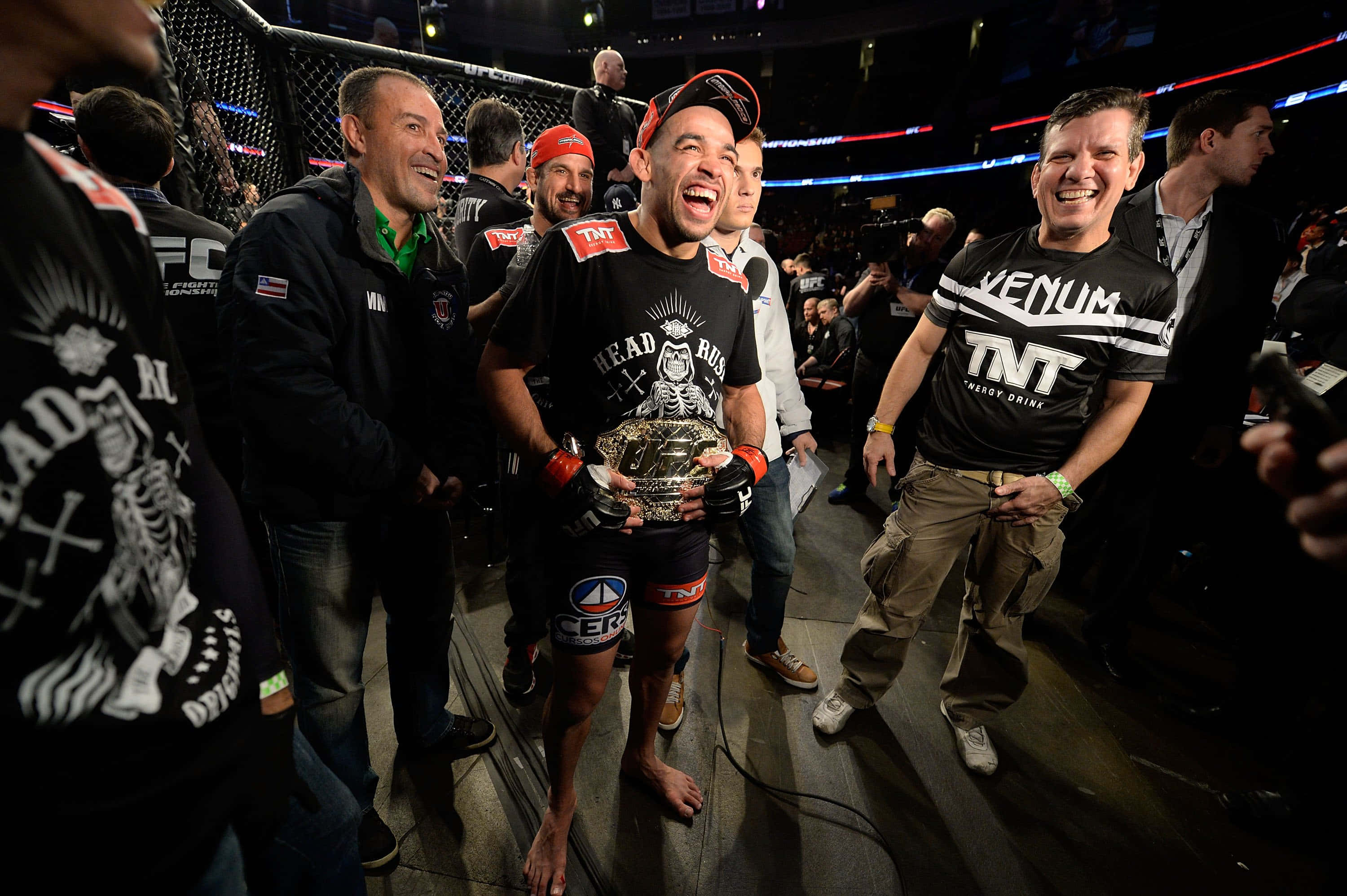 UFC 桌布 壁紙 — 一位勝利的MMA拳手在燈光明亮的八角籠中，被歡呼的團隊成員包圍，高興地笑著舉起冠軍腰帶。