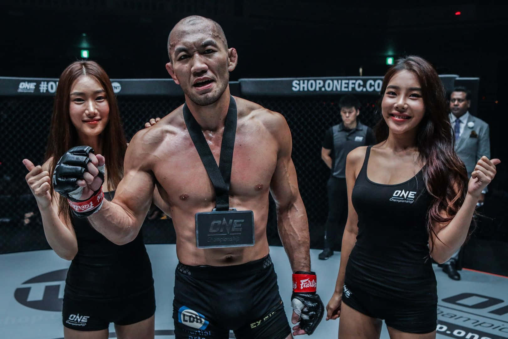 UFC 桌布 壁紙 — 一位勝利的MMA拳手赤裸上身站在擂台上，兩邊有兩位微笑的擂台女郎拿著他的手套。