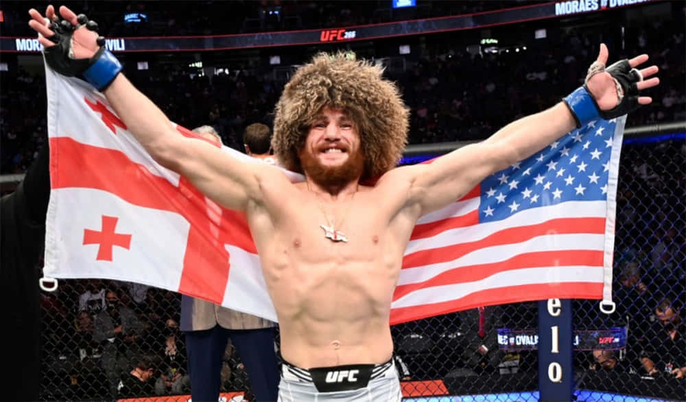 桌布 壁紙 — 一位勝利的UFC拳手，留著大捲鬍鬚並戴著毛帽，在八角籠中手持喬治亞和美國國旗慶祝。