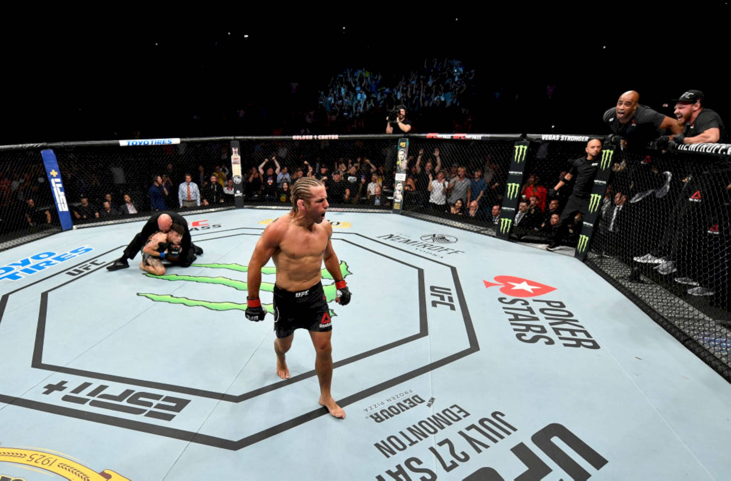 MMA 桌布 壁紙 — 一位勝利的UFC拳手在八角籠中威風凜凜地站立，而他的對手在明亮的場館燈光下由工作人員照料。