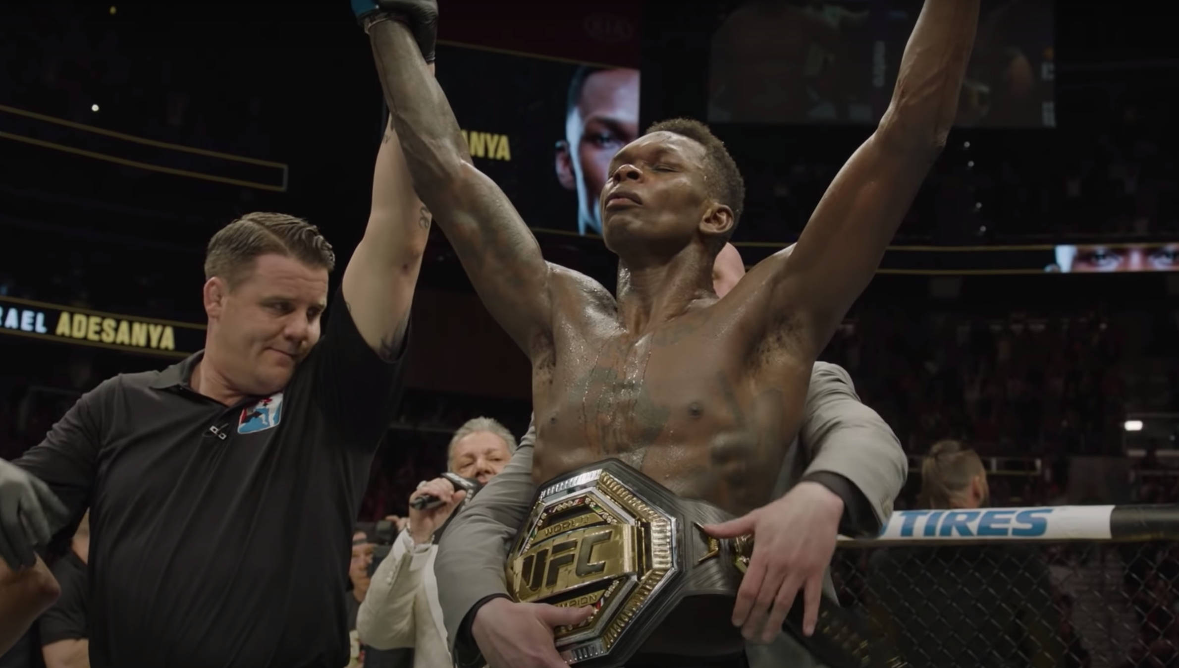 Adesanya 桌布 壁紙 — 一位勝利的UFC拳手在八角籠中高舉雙臂慶祝，裁判則舉起他的冠軍腰帶。