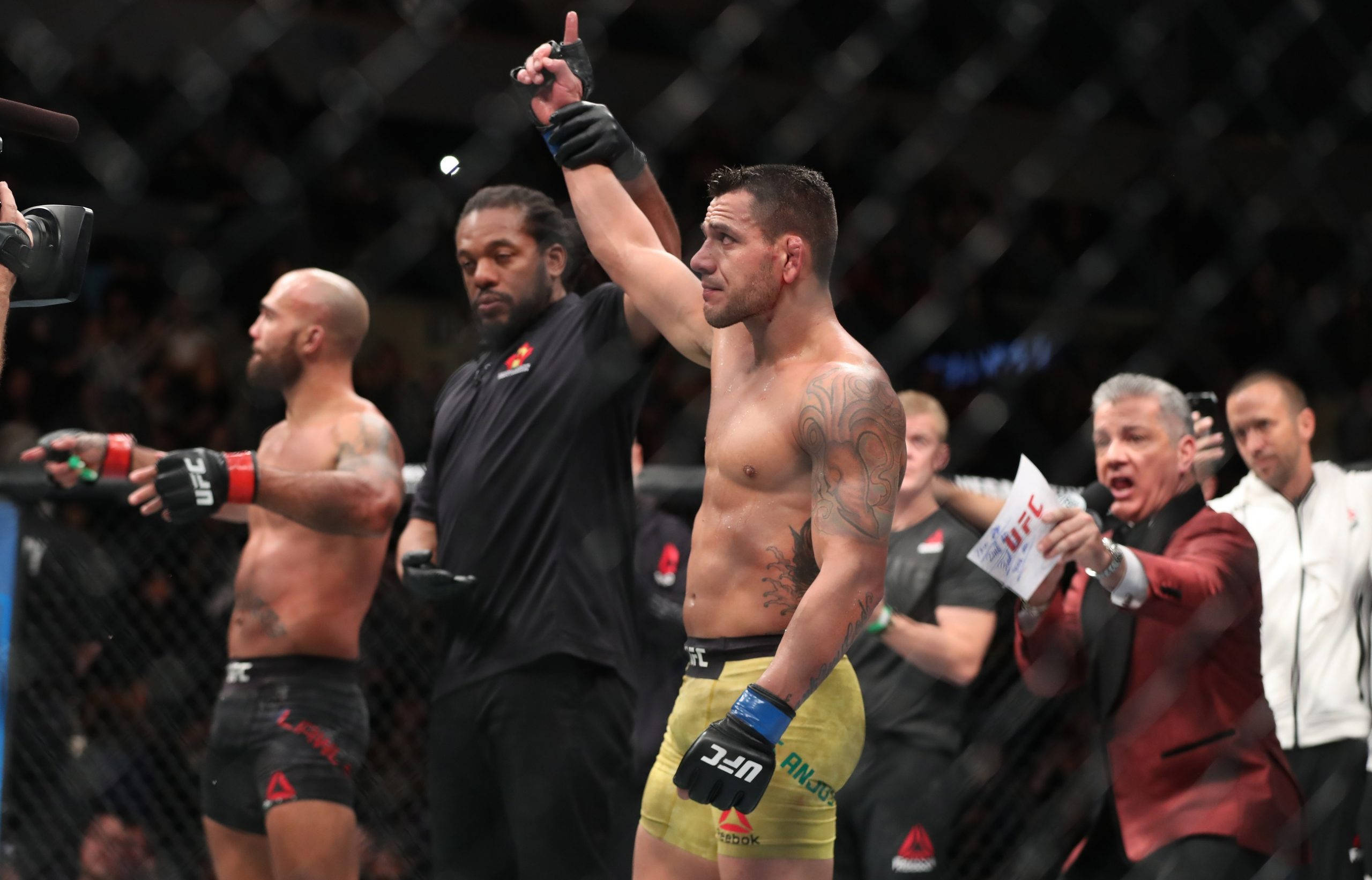 Victory 桌布 壁紙 — 一位勝利的UFC拳手在八角籠內舉起手指慶祝勝利，周圍的官員和評論員作出反應。