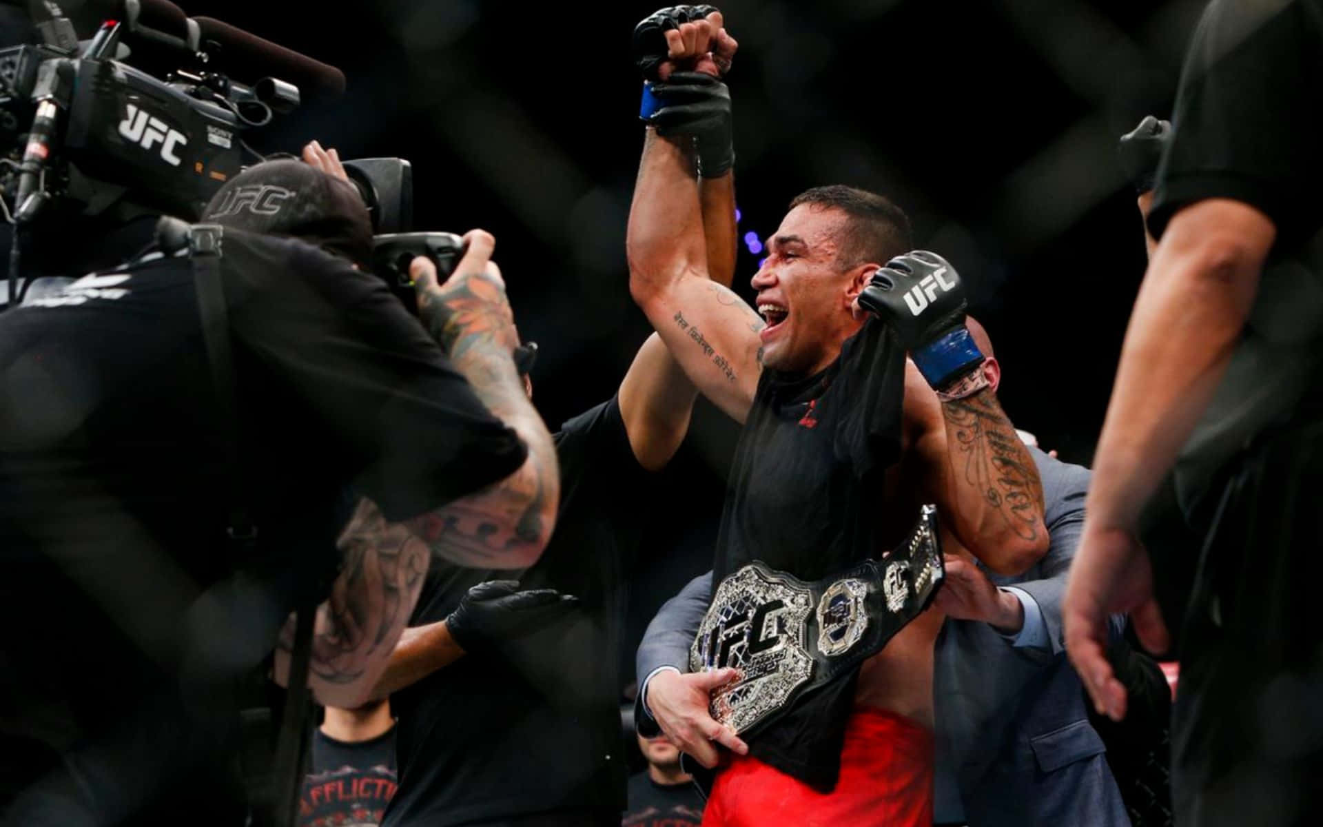 Athletic 桌布 壁紙 — 一位勝利的UFC拳手在八角籠內被攝影機和工作人員包圍，高舉冠軍腰帶和拳頭慶祝勝利。