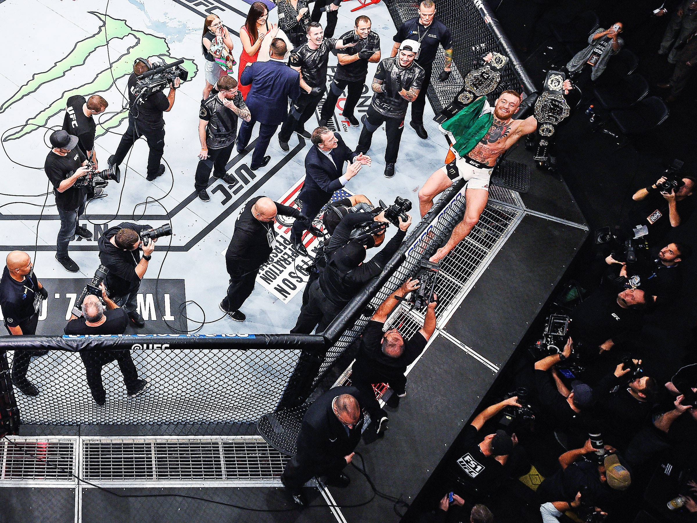 UFC 桌布 壁紙 — 一位勝利的UFC拳手在八角籠頂部慶祝，攝影師和官員圍繞著他，呈現出高能量的賽後時刻。