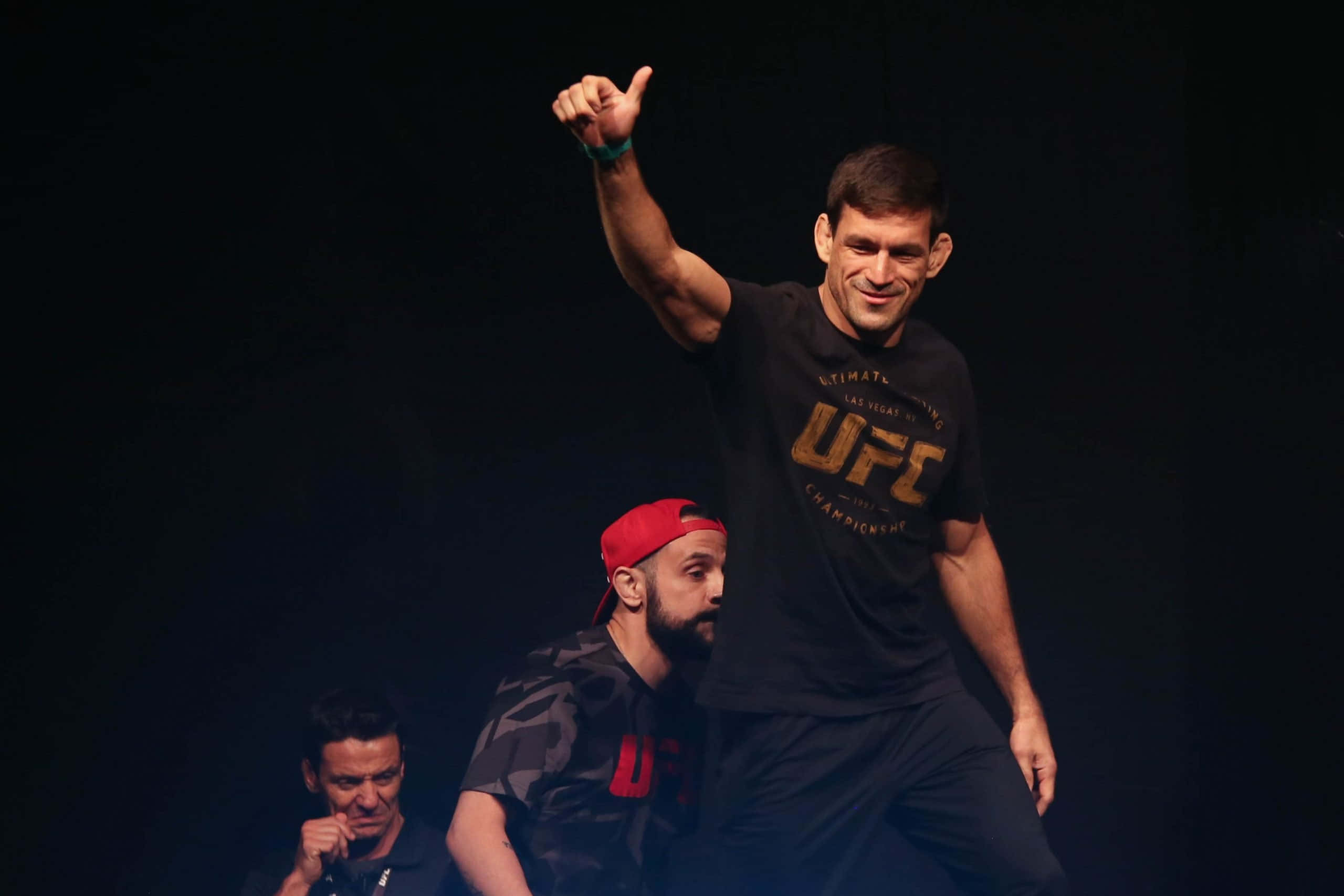 Champion 桌布 壁紙 — 一位勝利的UFC拳手在戲劇性的舞台燈光下豎起拇指慶祝勝利，身後有兩名男子。