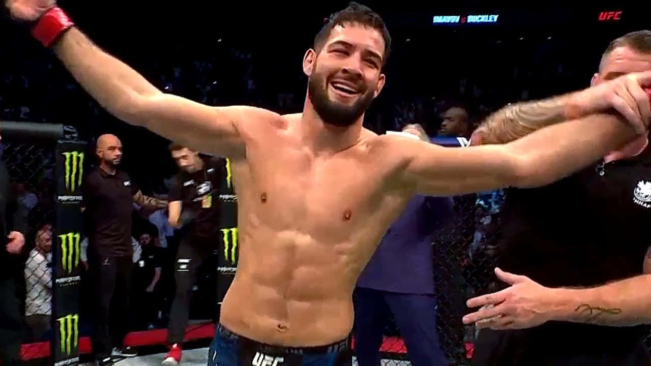 UFC 桌布 壁紙 — 一位勝利的UFC拳手在明亮的場館燈光下，在八角籠中高舉雙臂慶祝。