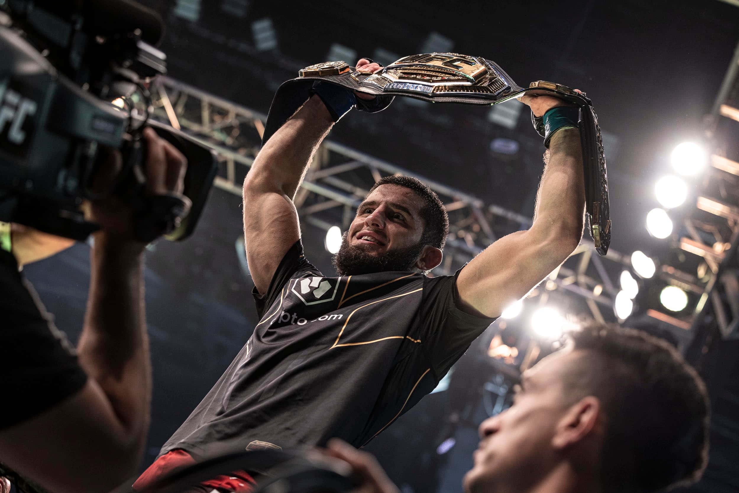 MMA 桌布 壁紙 — 一位勝利的UFC拳手在明亮的場館燈光下高舉冠軍腰帶，相機捕捉了這一刻。