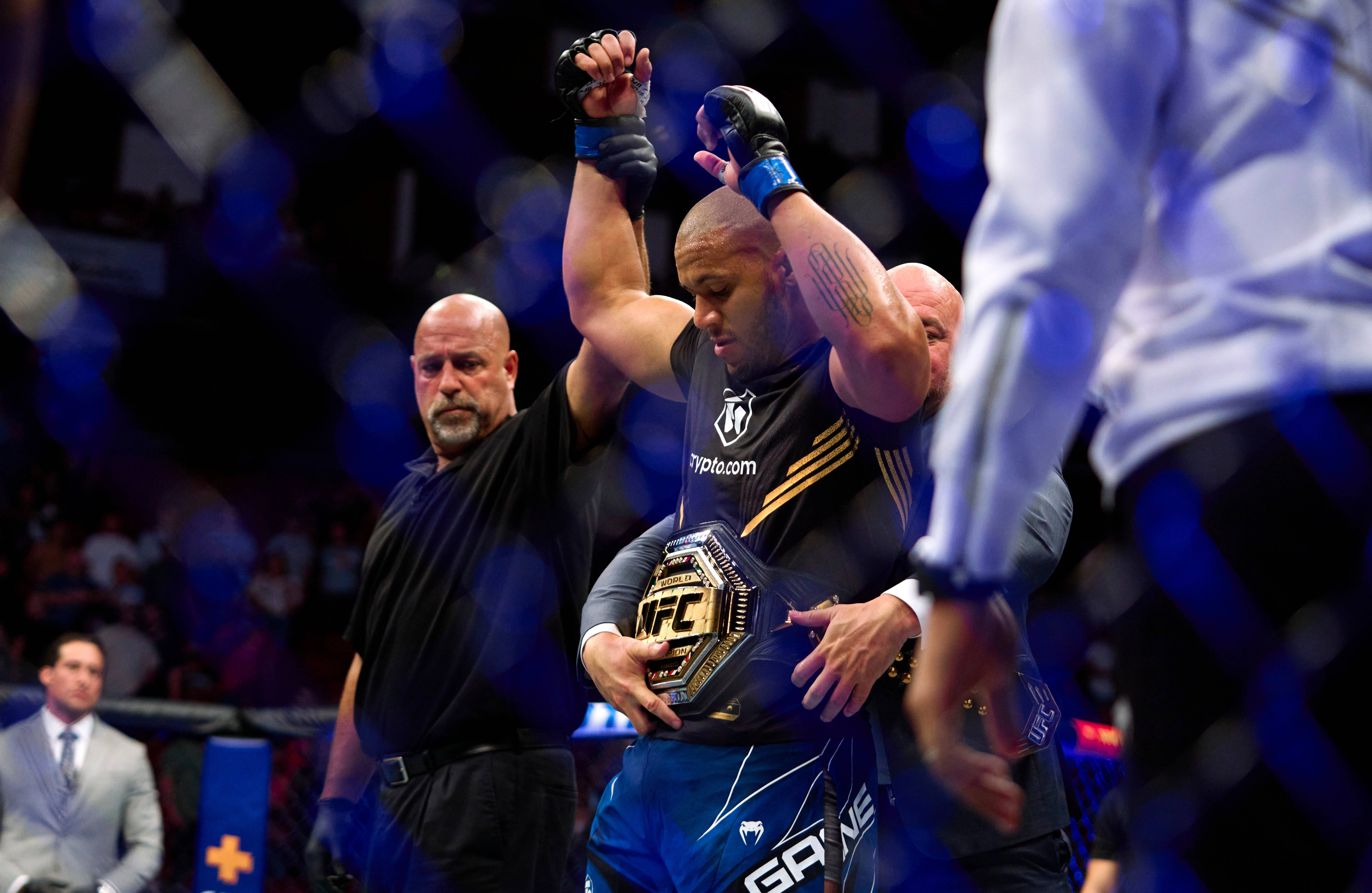 Victory 桌布 壁紙 — 一位勝利的UFC拳手在明亮的場館燈光下，於八角籠中高舉冠軍腰帶和手臂，裁判宣佈他為勝者。