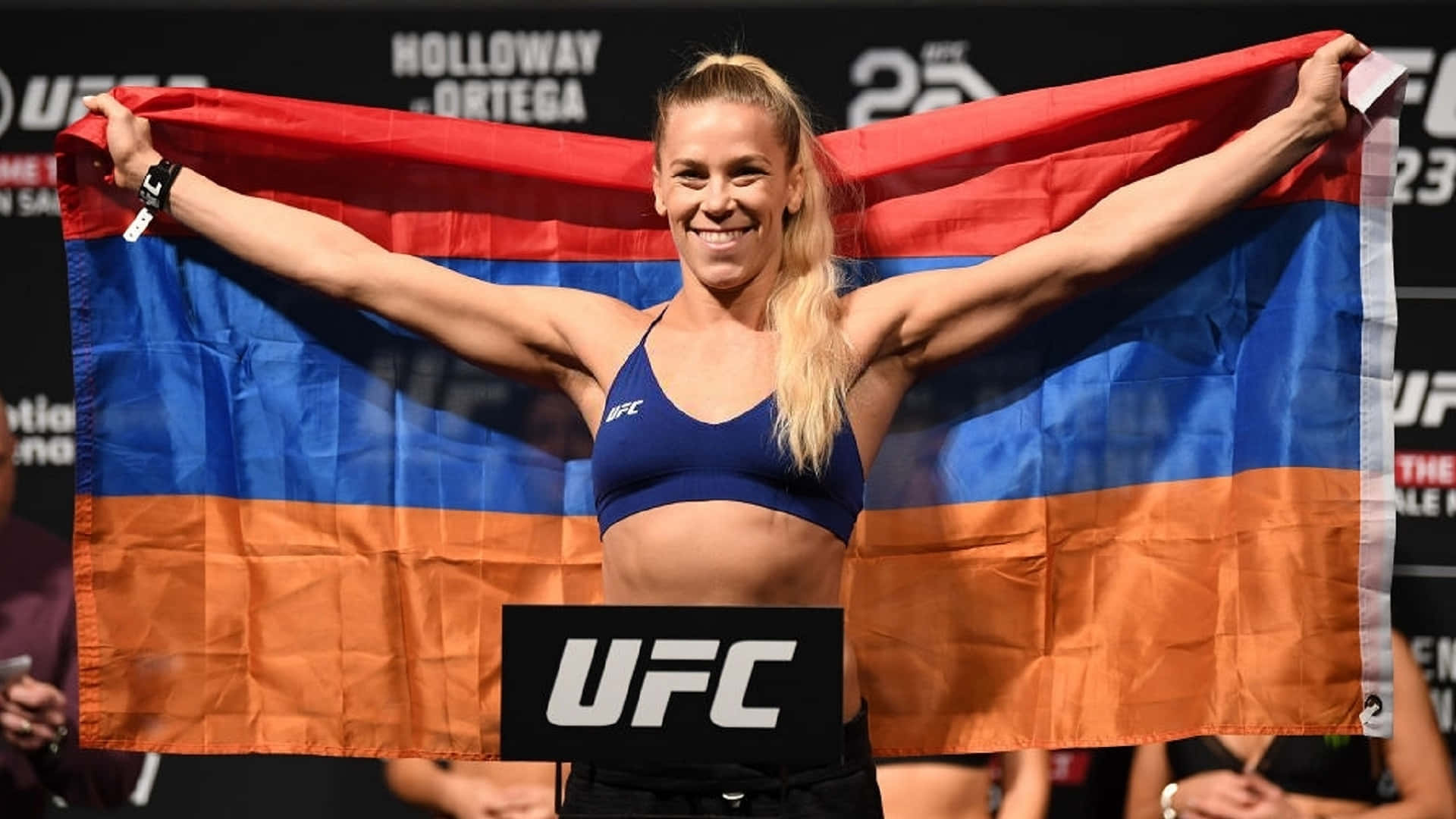 桌布 壁紙 — 一位勝利的UFC拳手在稱重活動中自豪地展開亞美尼亞國旗，滿臉光彩。