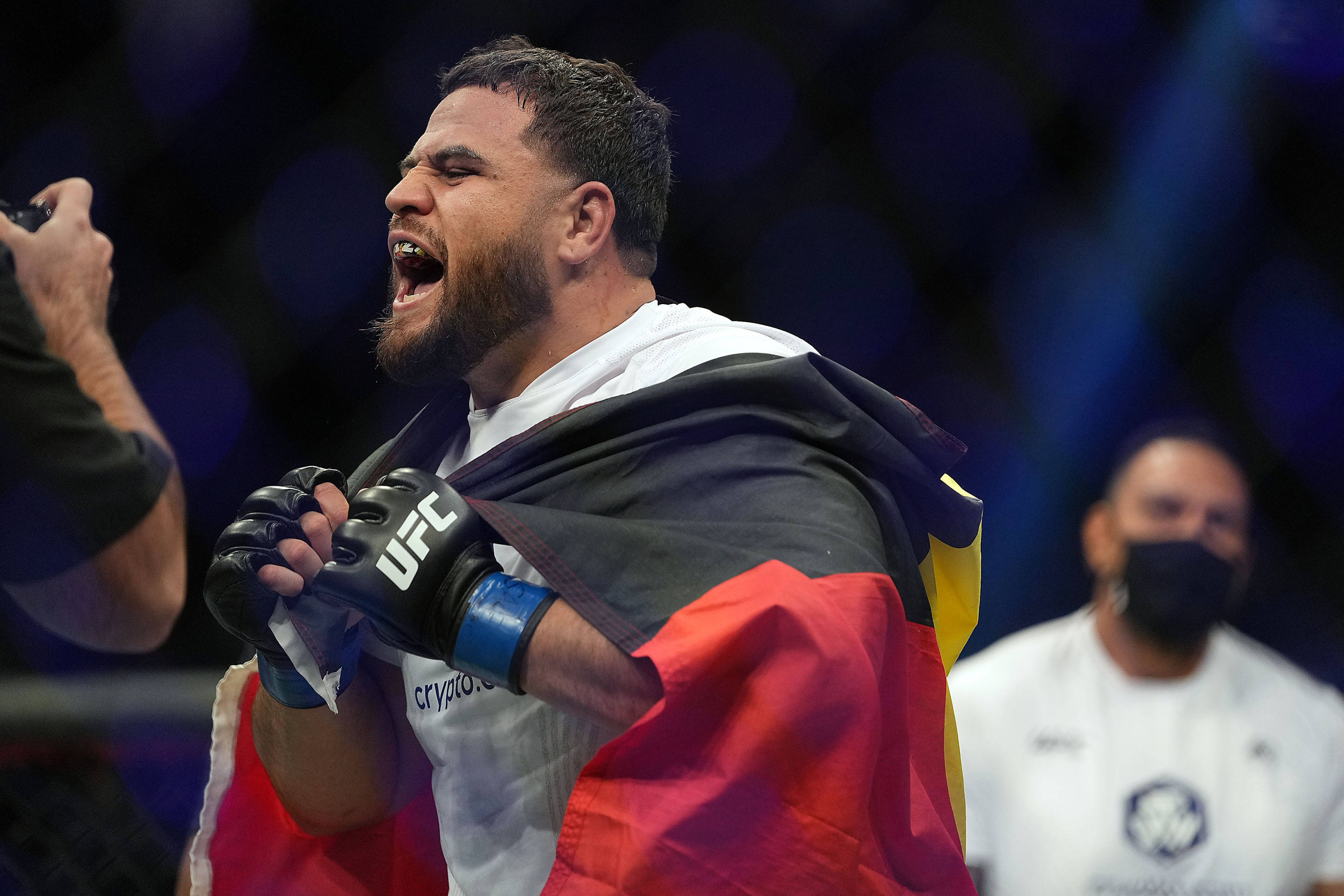 MMA 桌布 壁紙 — 一位勝利的UFC拳手披著德國國旗，在明亮的場館燈光下於八角籠內咆哮慶祝勝利。