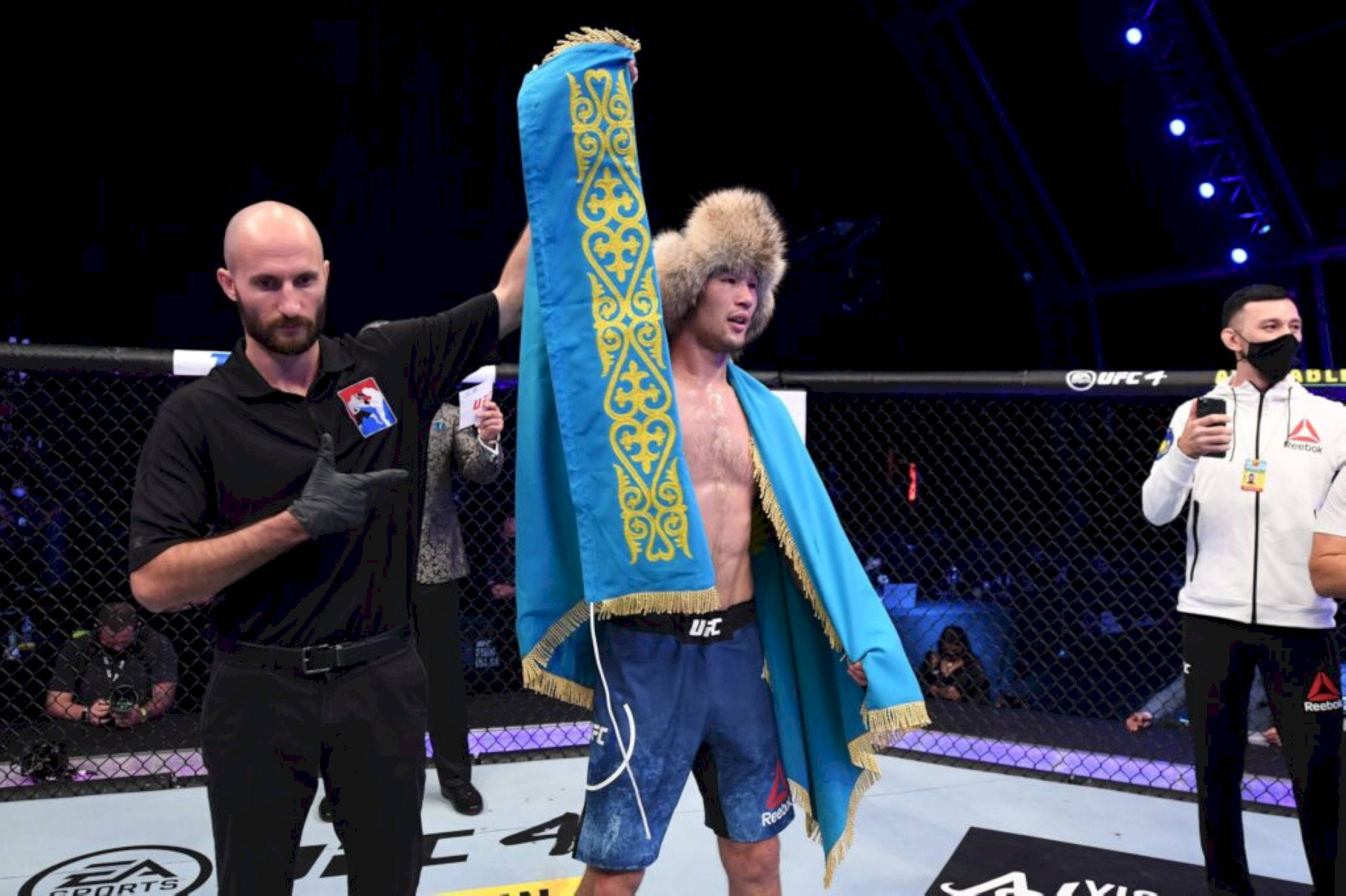 UFC 桌布 壁紙 — 一位勝利的UFC拳手披著藍金色華麗的哈薩克旗幟，站在八角籠中，裁判舉起他的手。