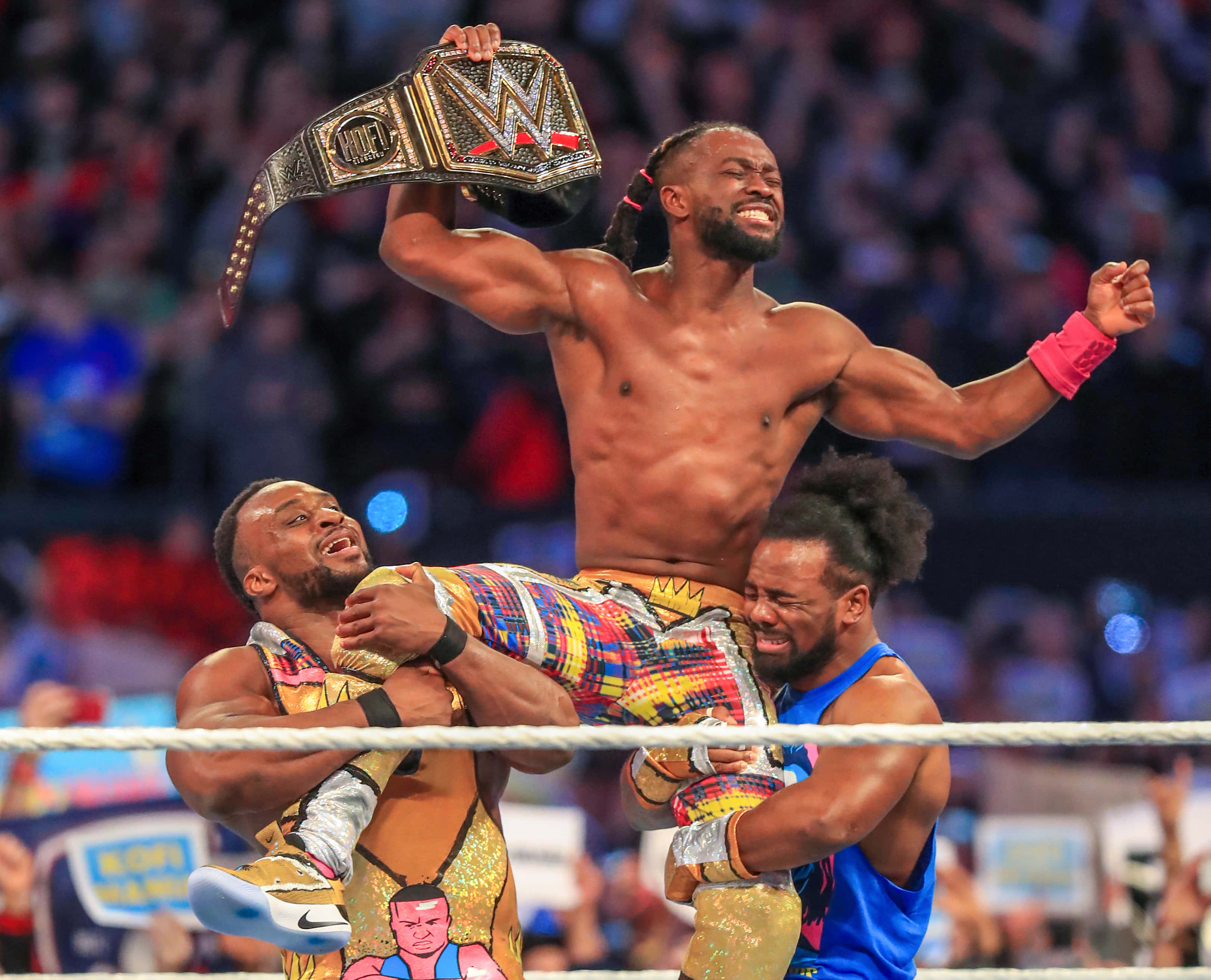 WWE 桌布 壁紙 — 一位勝利的WWE摔角手高舉冠軍腰帶，同時被兩名對手在摔角擂台上抬起。
