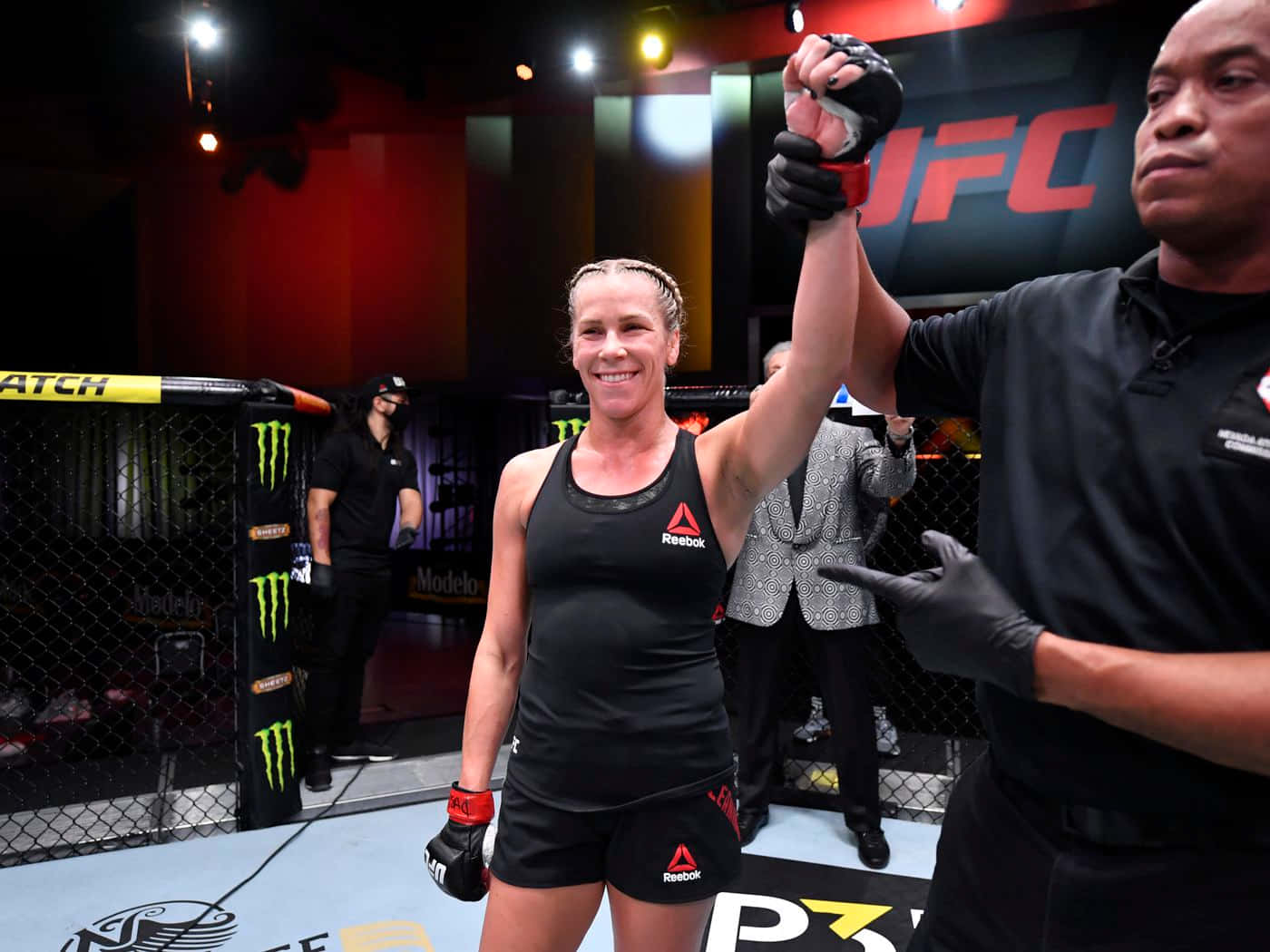 桌布 壁紙 — 一位勝利的女性MMA拳手在明亮的場館燈光下，於UFC八角籠內被裁判舉起手時微笑。