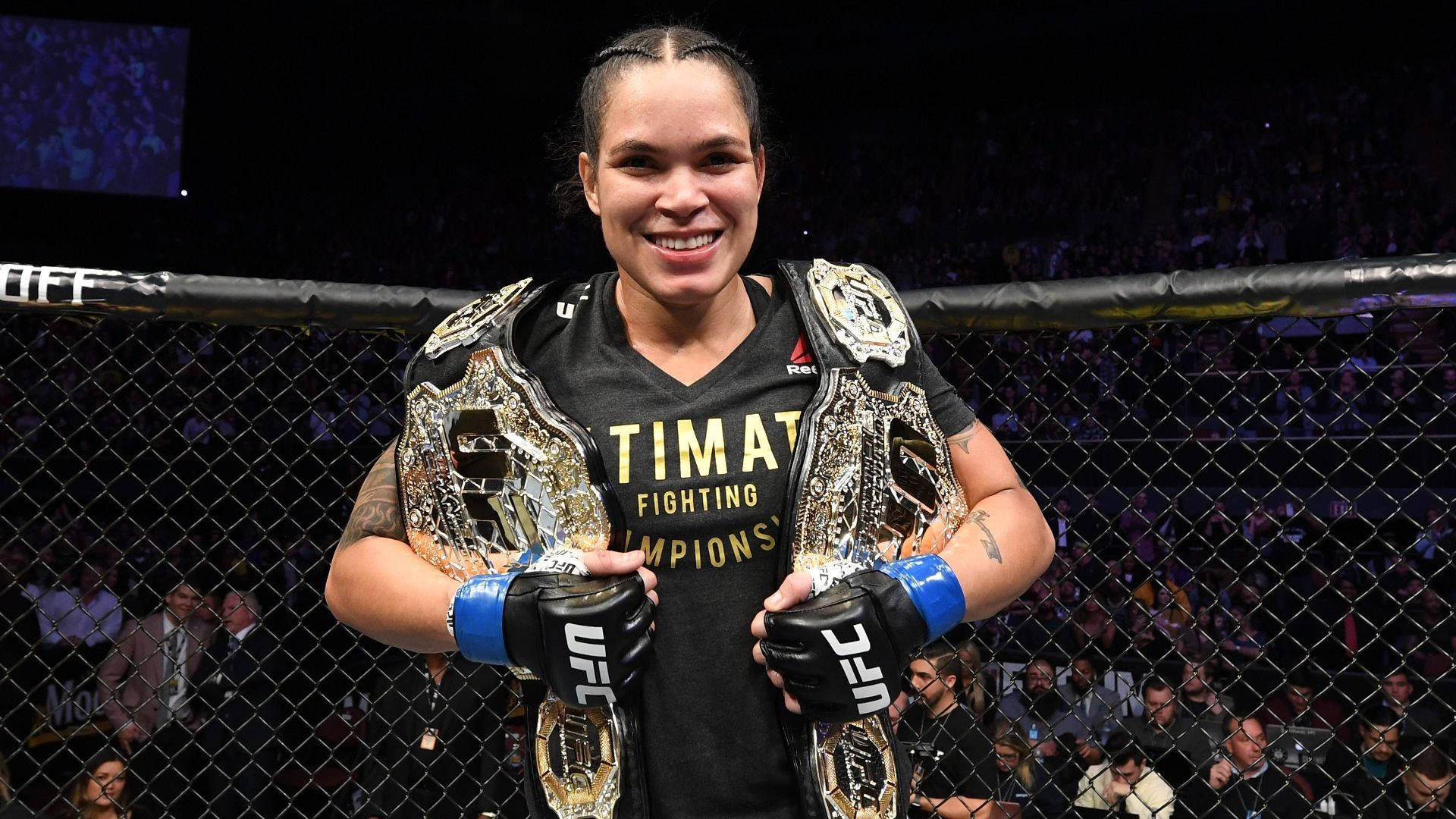 Champion 桌布 壁紙 — 一位勝利的女性UFC拳手在八角籠內、場館燈光下，手持兩條冠軍腰帶，笑容燦爛。