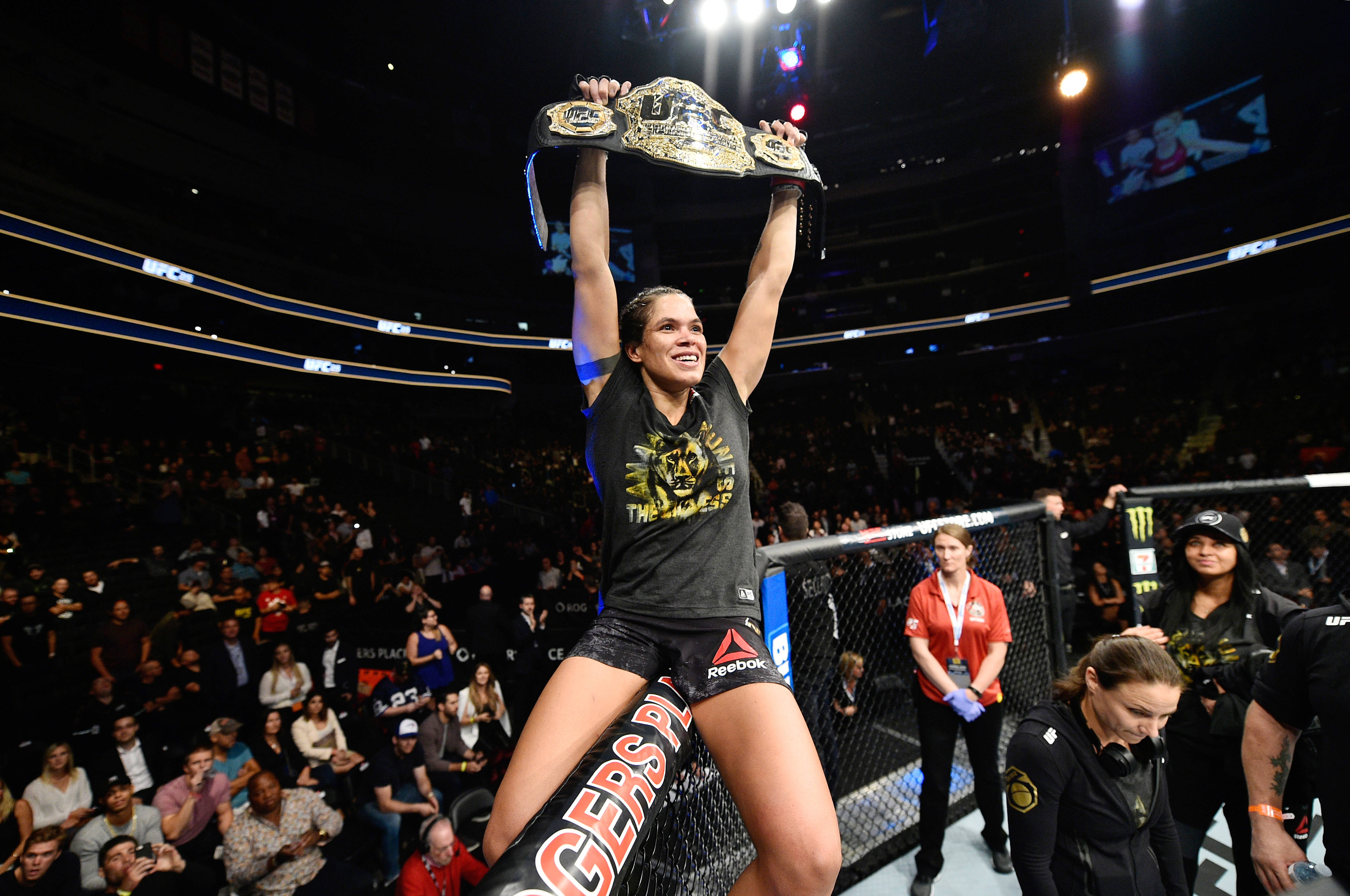Champion 桌布 壁紙 — 一位勝利的女性拳手在燈光明亮的八角籠內，將冠軍腰帶高舉過頭慶祝勝利。