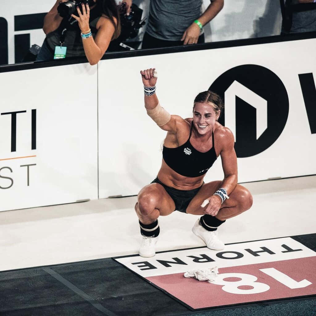 Athletic 桌布 壁紙 — 一位勝利的女性運動員蹲在比賽墊上，在明亮的場館燈光下舉起拳頭慶祝勝利。
