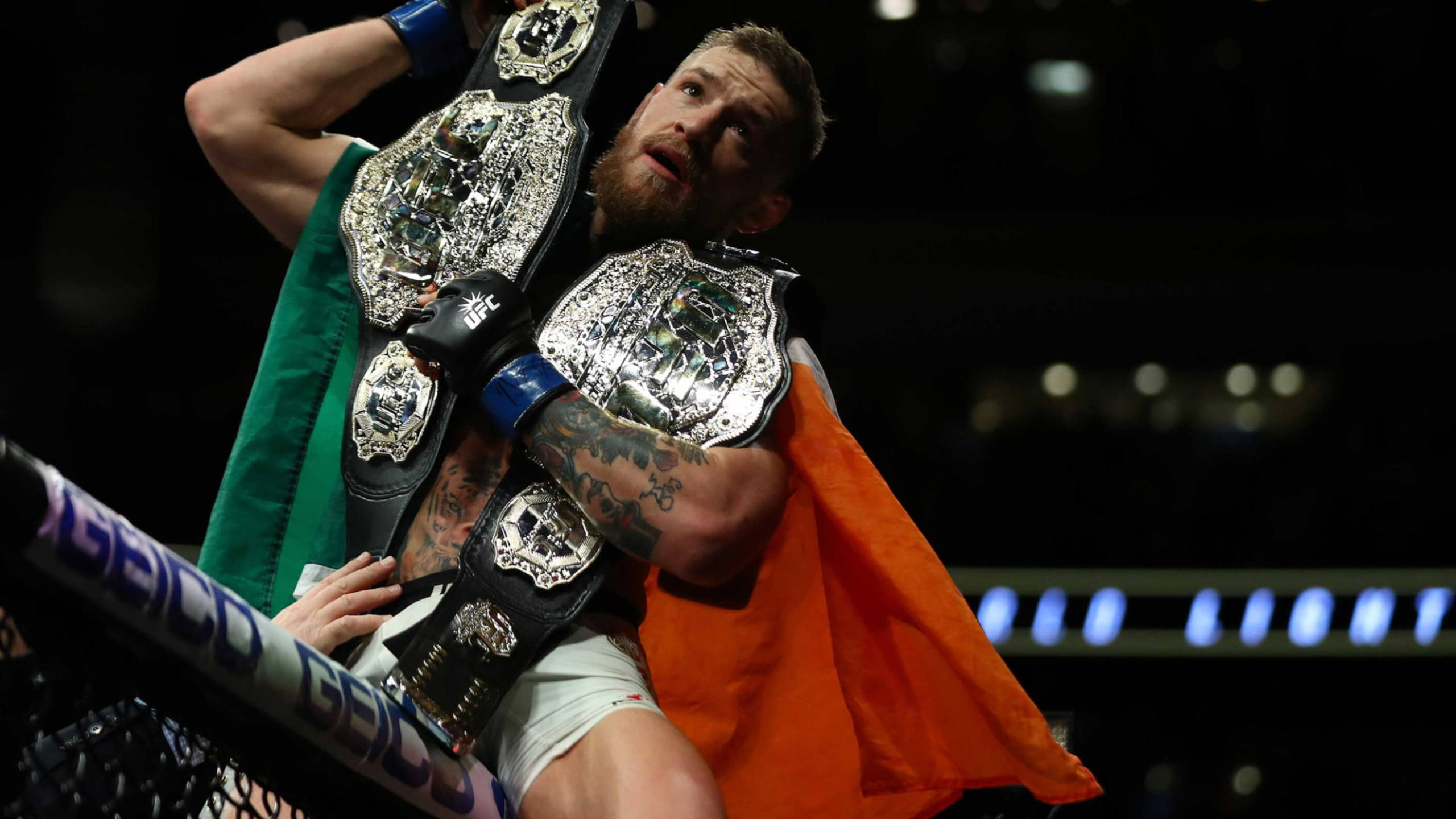 Mcgregor 桌布 壁紙 — 一位勝利的戰士披著綠色和橙色旗幟，在燈光明亮的競技場中高舉多條冠軍腰帶。