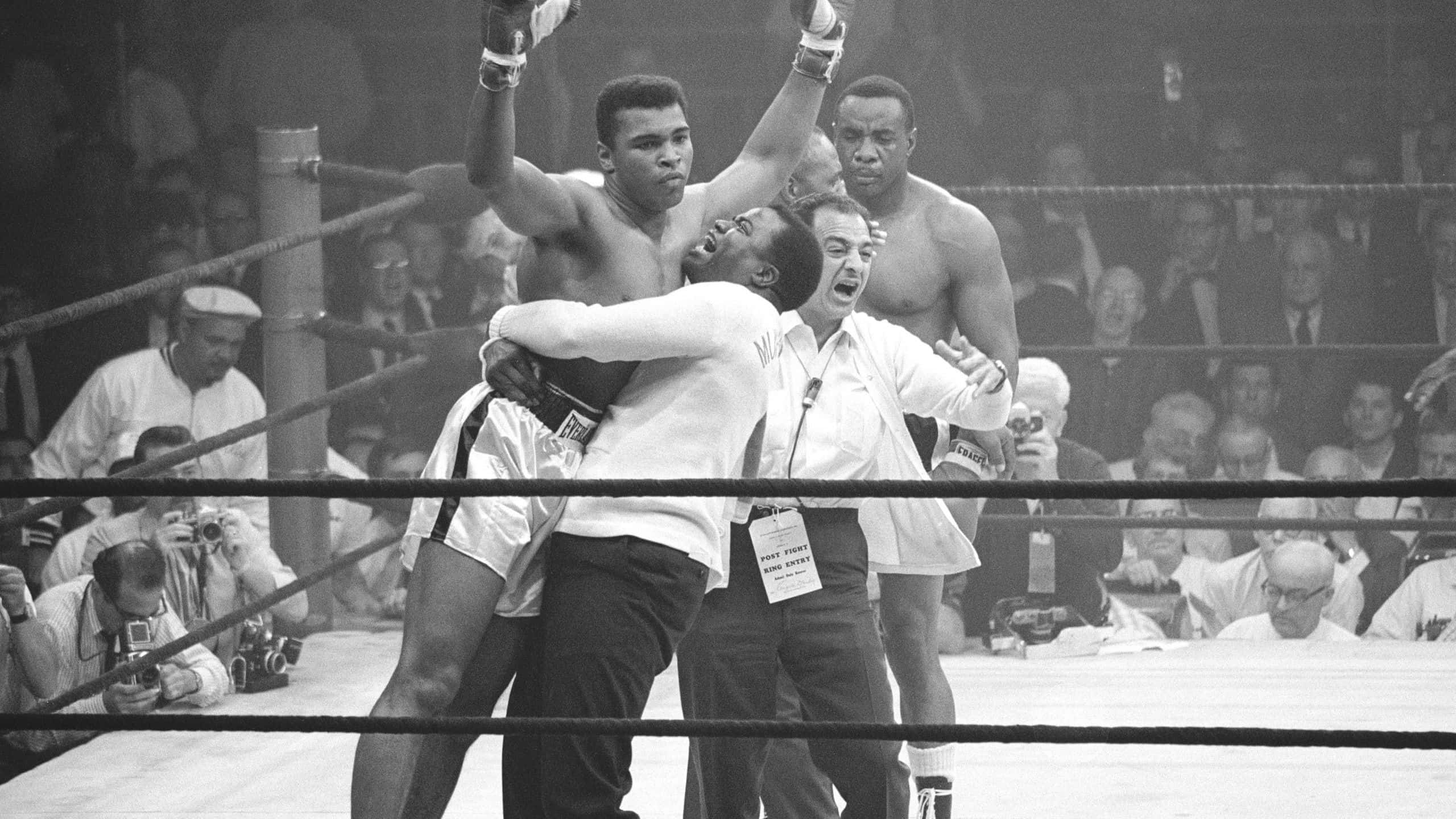 Boxing 桌布 壁紙 — 一位勝利的拳擊手在拳擊台上被教練舉起，裁判舉起他的手臂宣佈勝利。