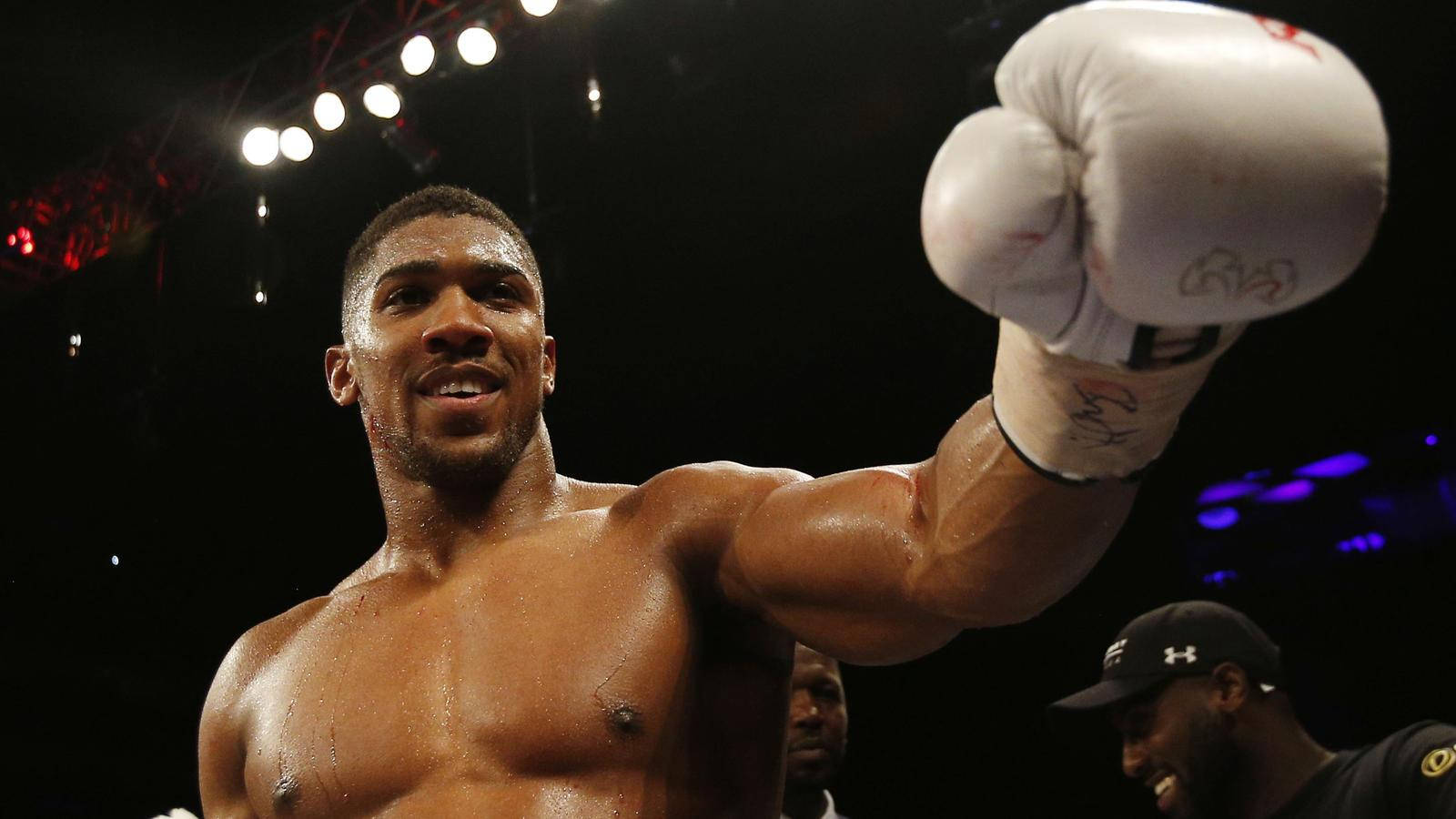 Anthony Joshua 桌布 壁紙 — 一位勝利的拳擊手在明亮的競技場燈光下，汗水在他肌肉發達的軀幹上閃閃發光，舉起戴著手套的拳頭慶祝勝利。