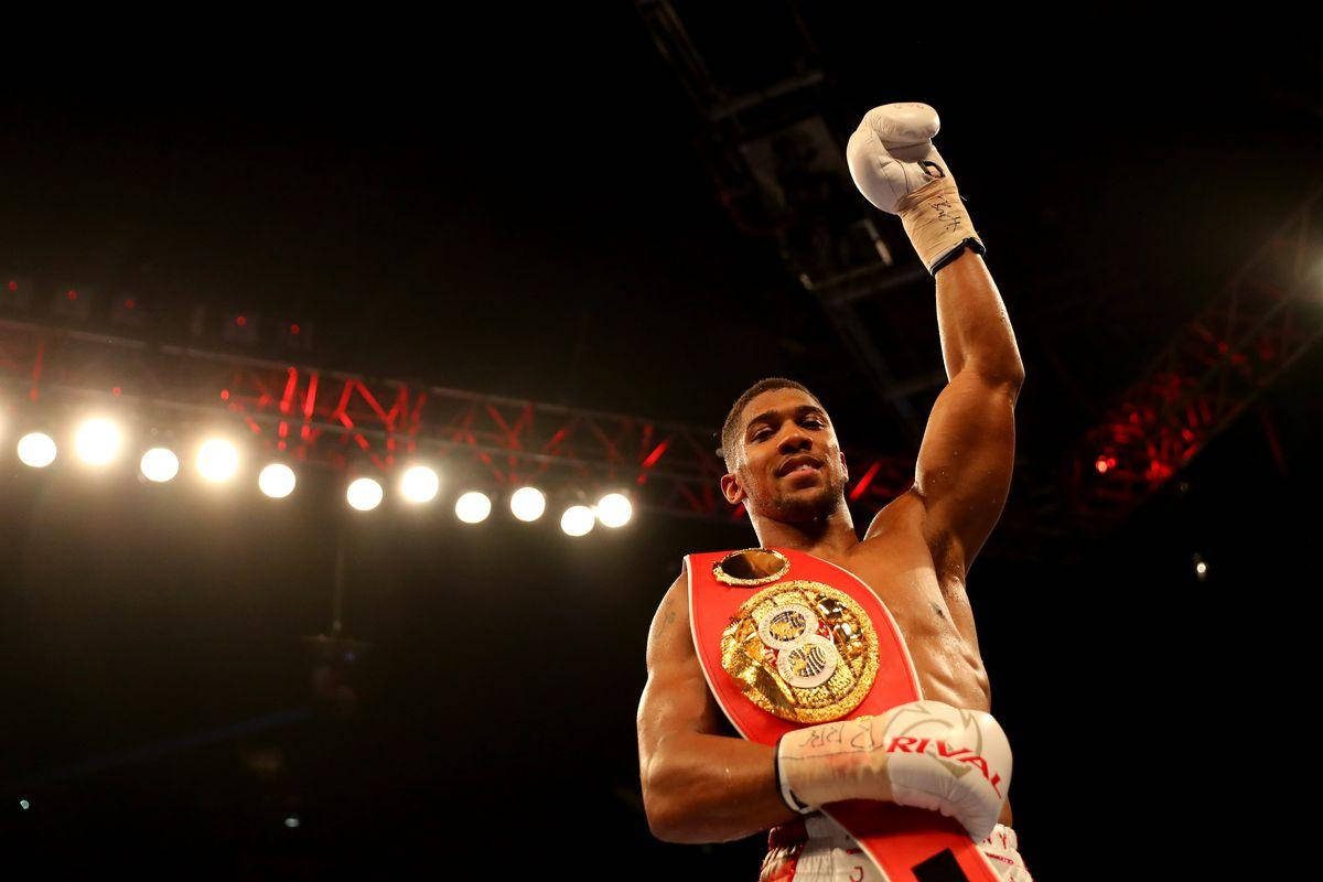 Anthony Joshua 桌布 壁紙 — 一位勝利的拳擊手在明亮的競技場燈光下舉起戴著手套的拳頭，胸前披著冠軍腰帶。
