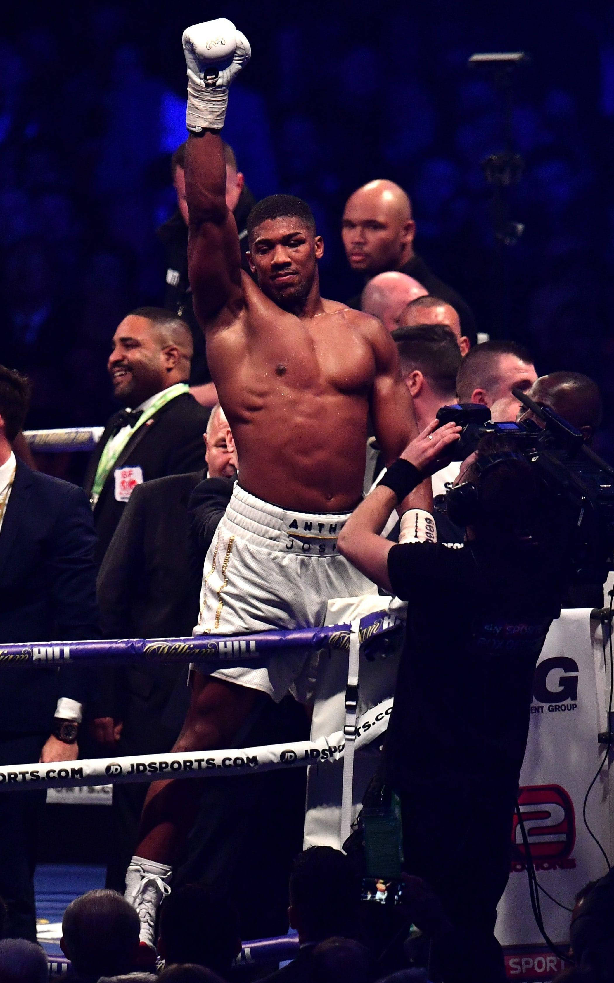 Anthony Joshua 桌布 壁紙 — 一位勝利的拳擊手在燈光明亮的擂台上，在歡呼的人群和攝影機的包圍下，舉起戴著手套的拳頭慶祝勝利。