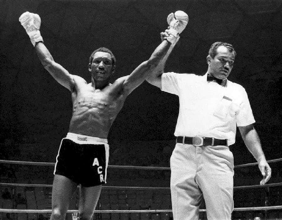 Champion 桌布 壁紙 — 一位勝利的拳擊手在燈光昏暗的擂台上高舉雙臂，裁判舉起他戴著手套的手宣告勝利。