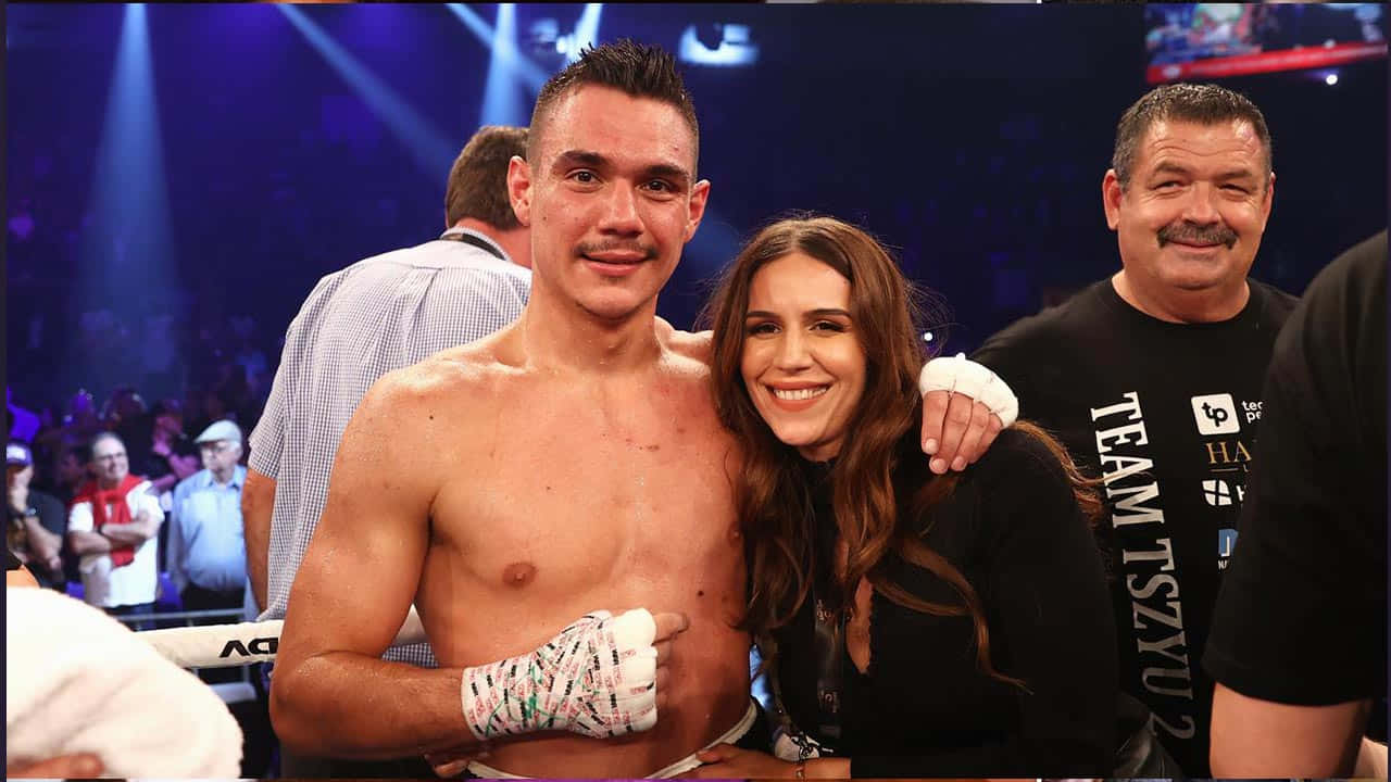 Tim Tszyu 桌布 壁紙 — 一位勝利的拳擊手纏著繃帶的手，在競技場燈光下與歡呼的伴侶和教練一起微笑。