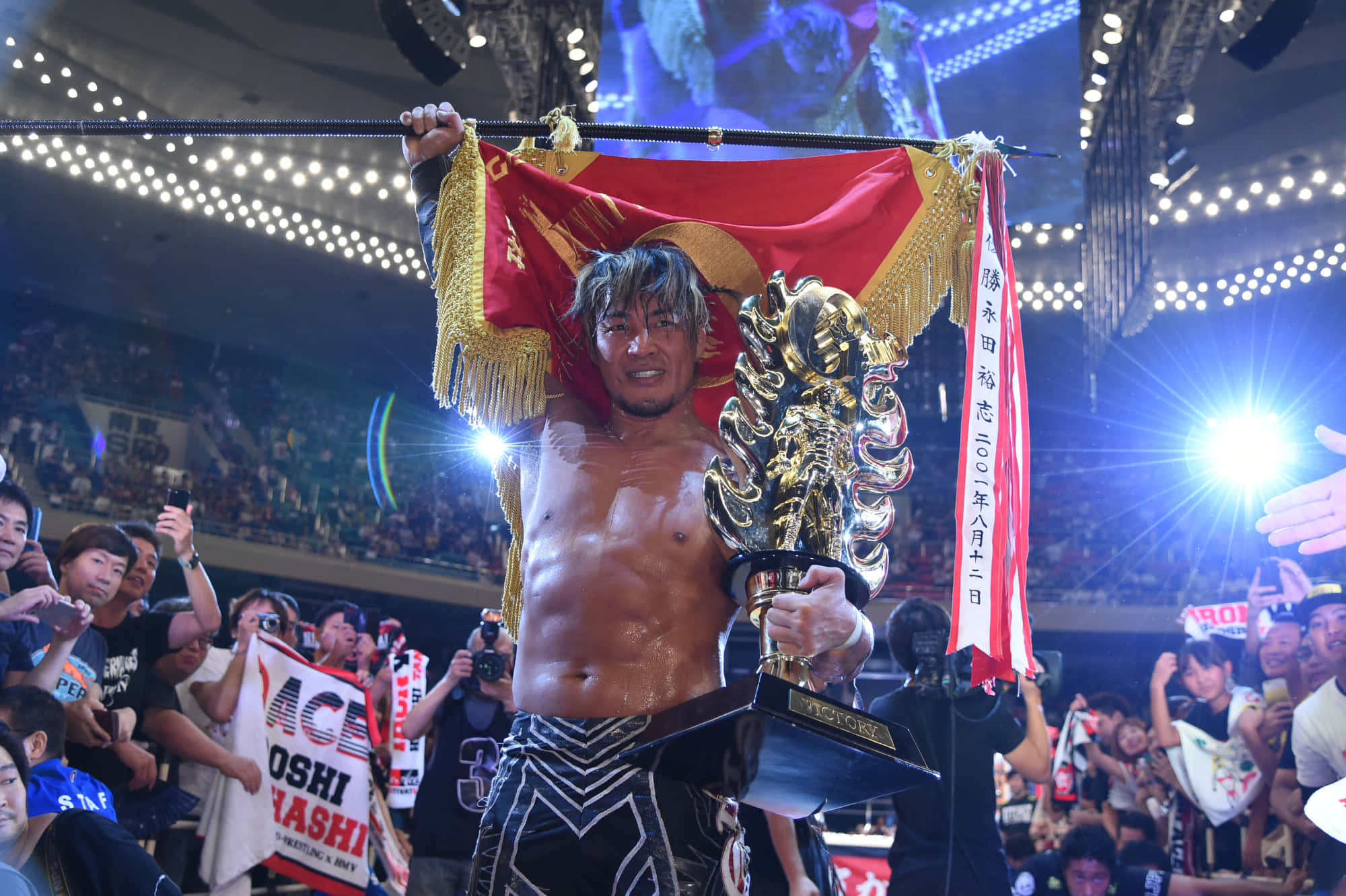 Champion 桌布 壁紙 — 一位勝利的摔角手在擠滿觀眾的場館中高舉冠軍獎盃和紅色旗幟慶祝勝利。