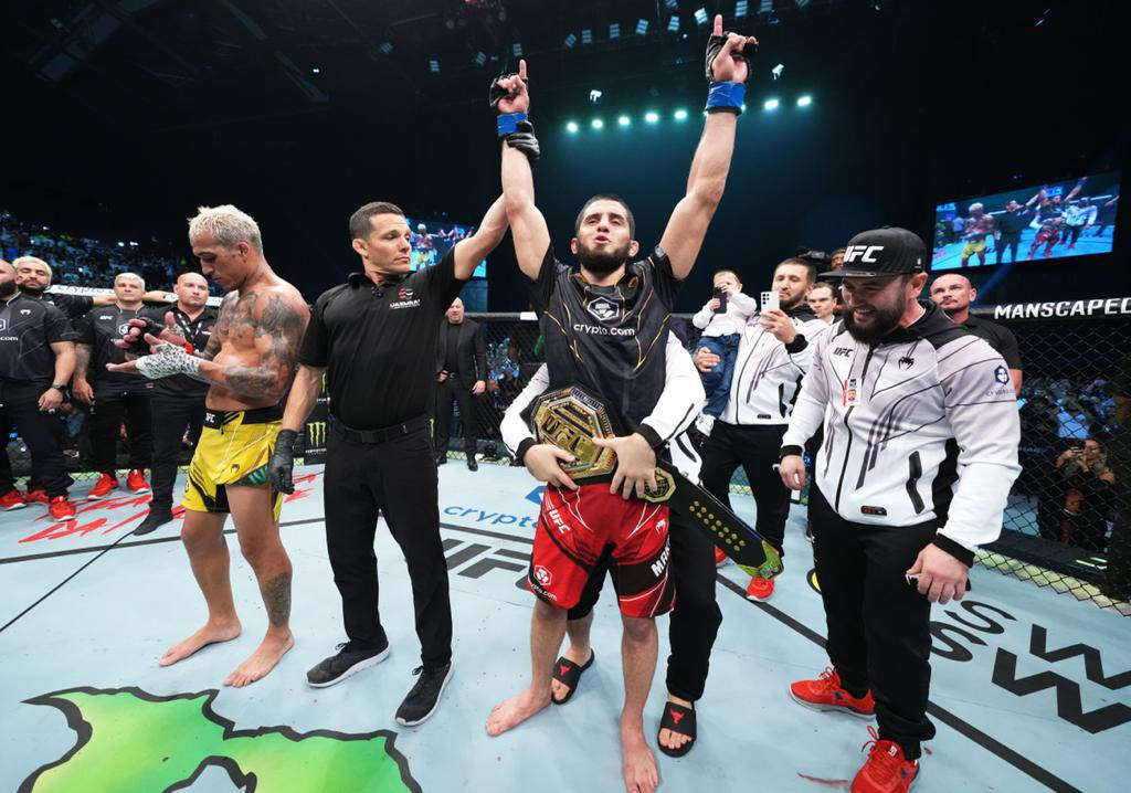 Champion 桌布 壁紙 — 一位勝利的混合武術家在八角籠中慶祝，手持冠軍腰帶，裁判舉起他的手。