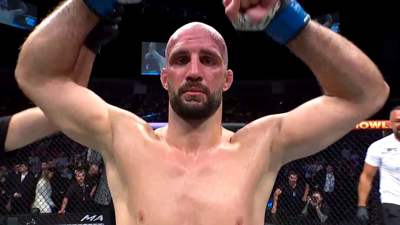 MMA 桌布 壁紙 — 一位勝利的混合武術家在燈光明亮的八角籠內高舉拳頭慶祝勝利，圍欄後是歡呼的人群。