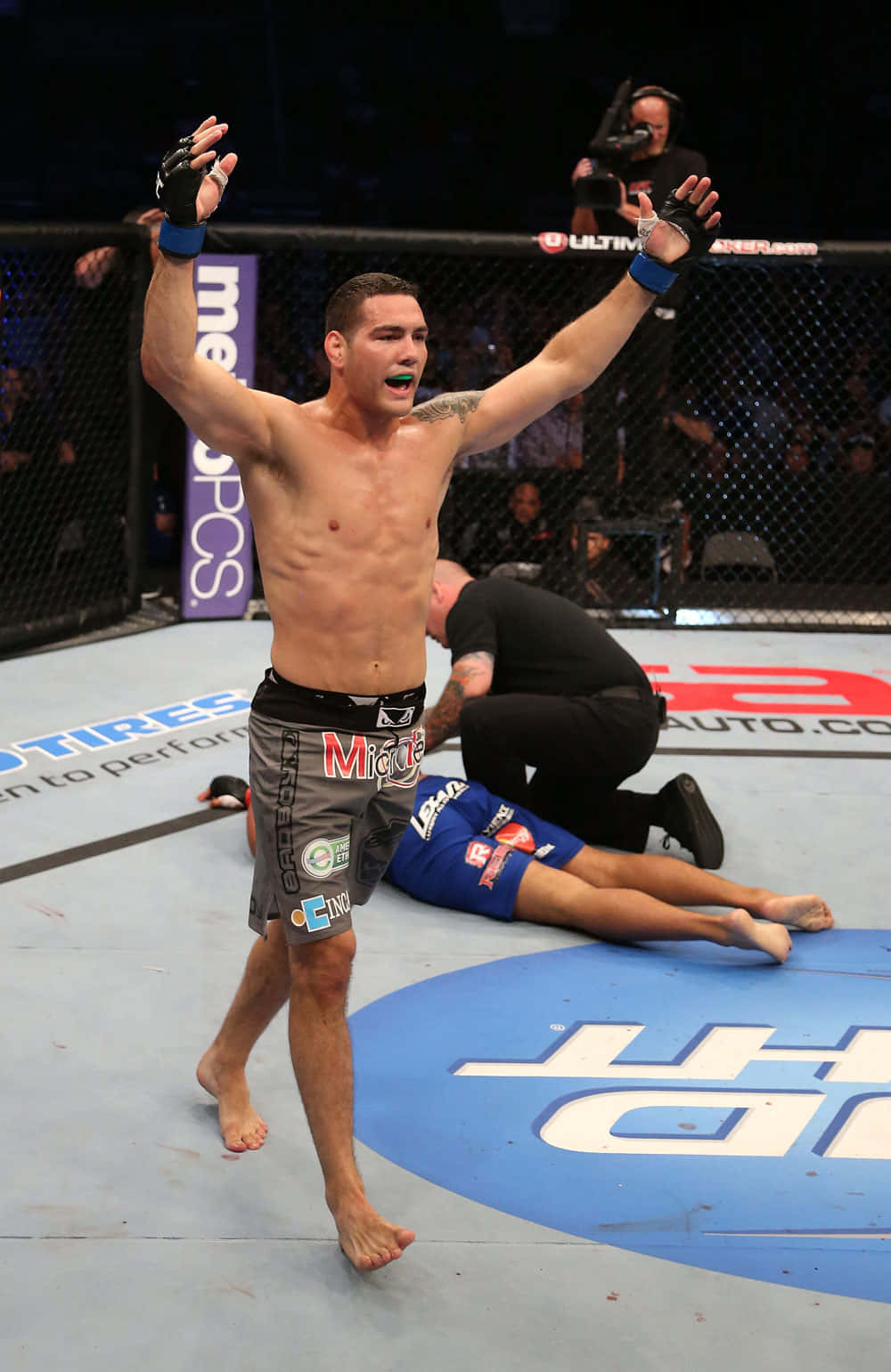 MMA 桌布 壁紙 — 一位勝利的混合武術家在籠內高舉雙臂慶祝勝利，而他的對手則倒在墊子上。
