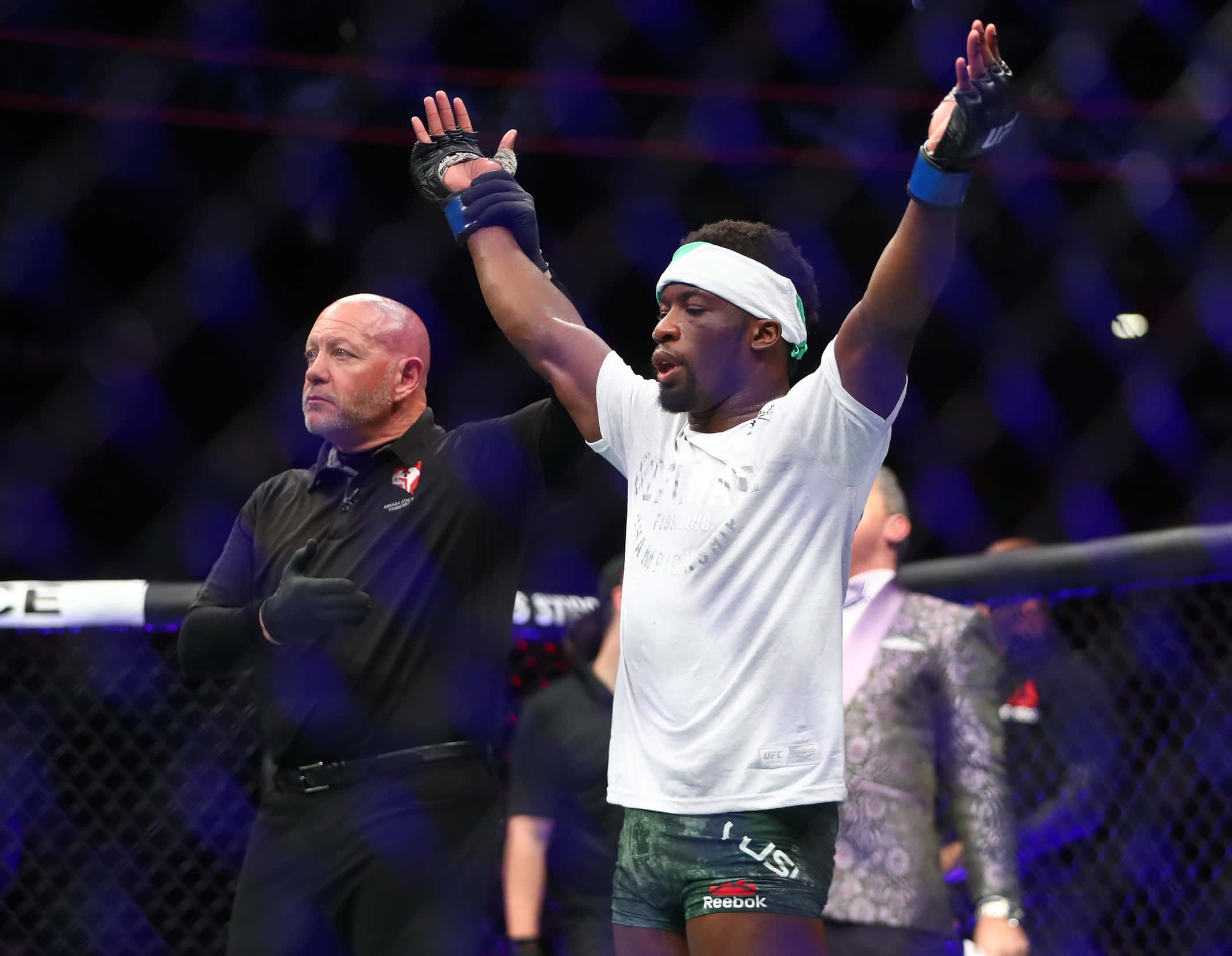MMA 桌布 壁紙 — 一位勝利的混合武術選手在燈光明亮的八角籠內高舉雙臂慶祝勝利，裁判示意他獲勝。