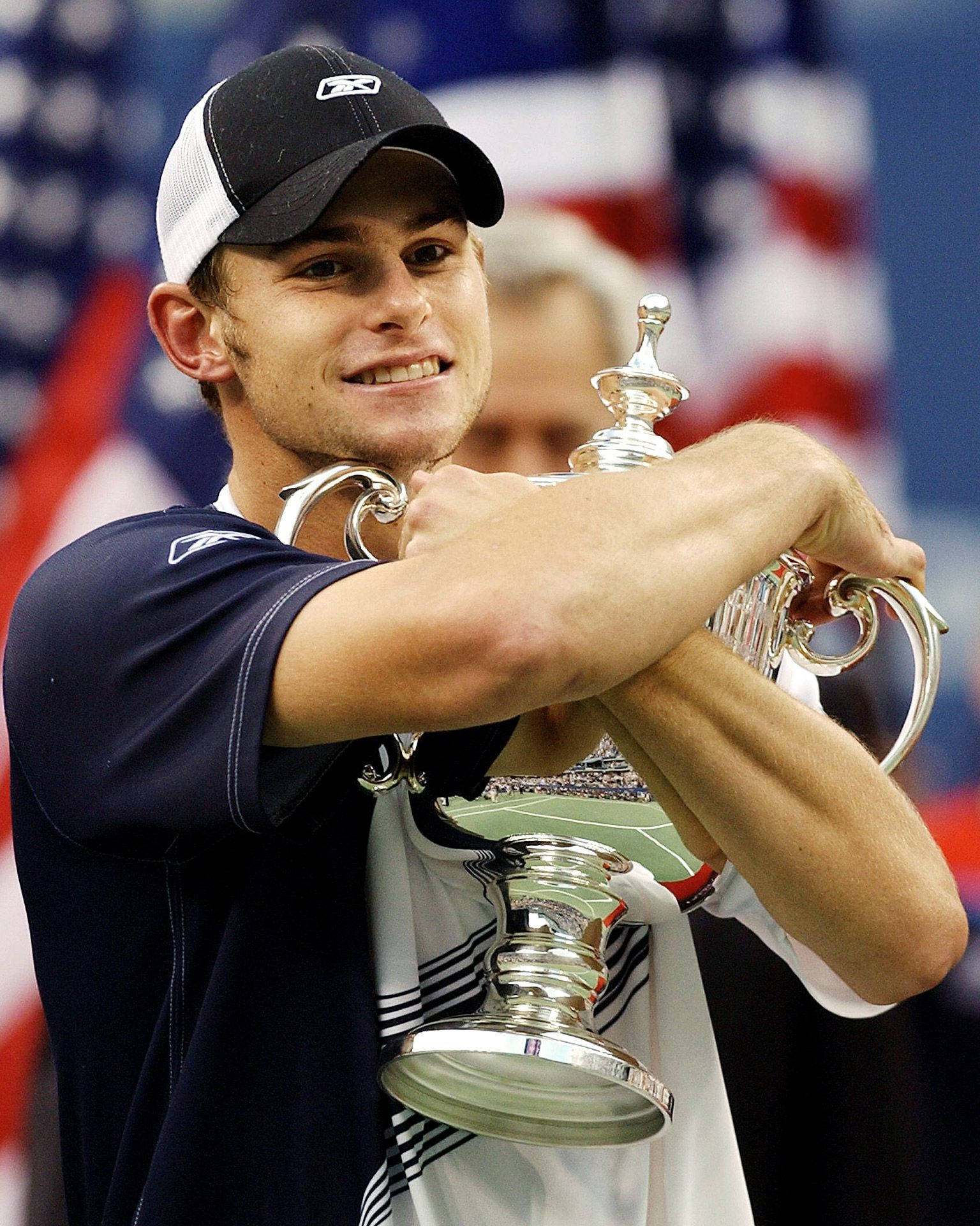 Trophy 桌布 壁紙 — 一位勝利的男性網球選手笑容滿面，手持一個大型銀色獎盃，背景是模糊的美國國旗。
