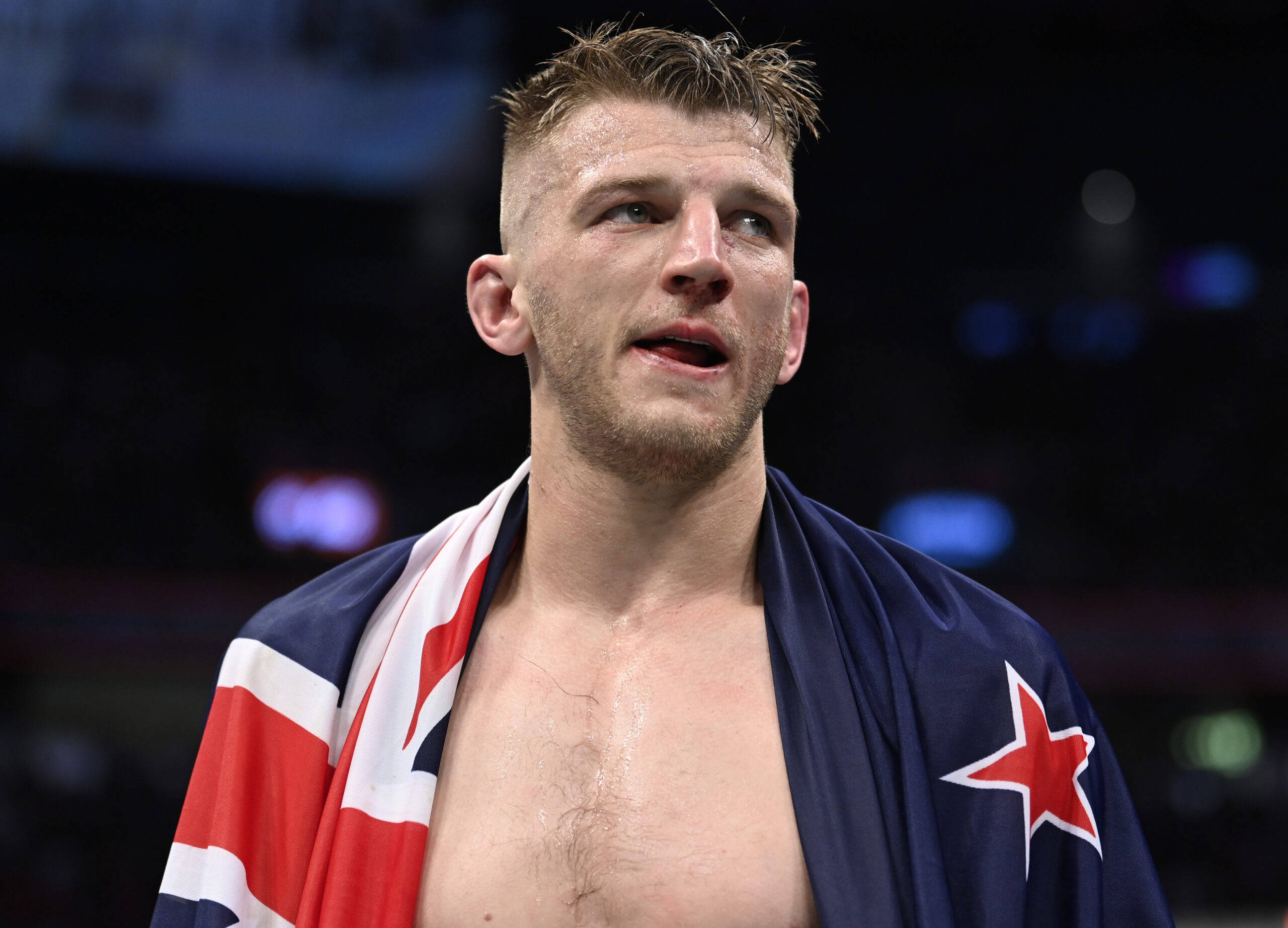 UFC 桌布 壁紙 — 一位勝利的男性運動員在燈光明亮的競技場中，披著英國國旗和紐西蘭國旗慶祝勝利。