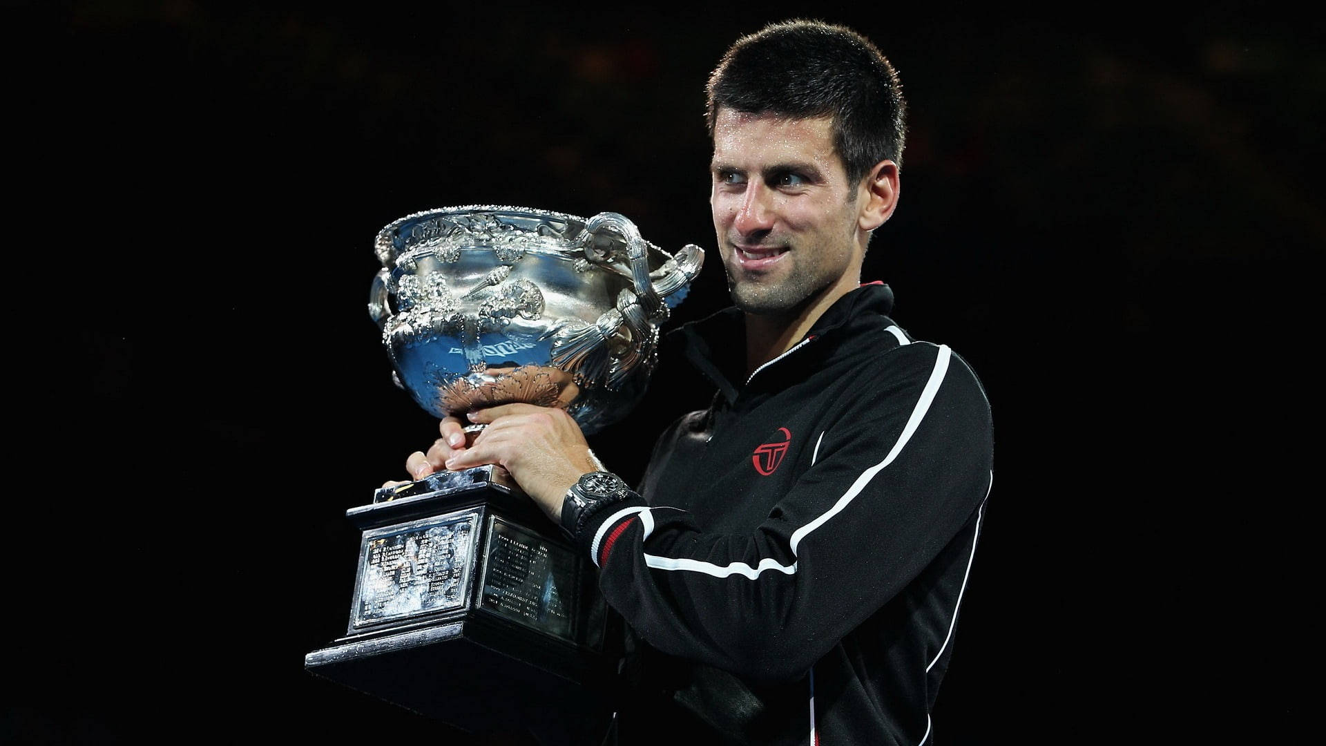 Novak 桌布 壁紙 — 一位勝利的網球運動員在黑暗背景下微笑著手持一個華麗的大銀色獎盃。