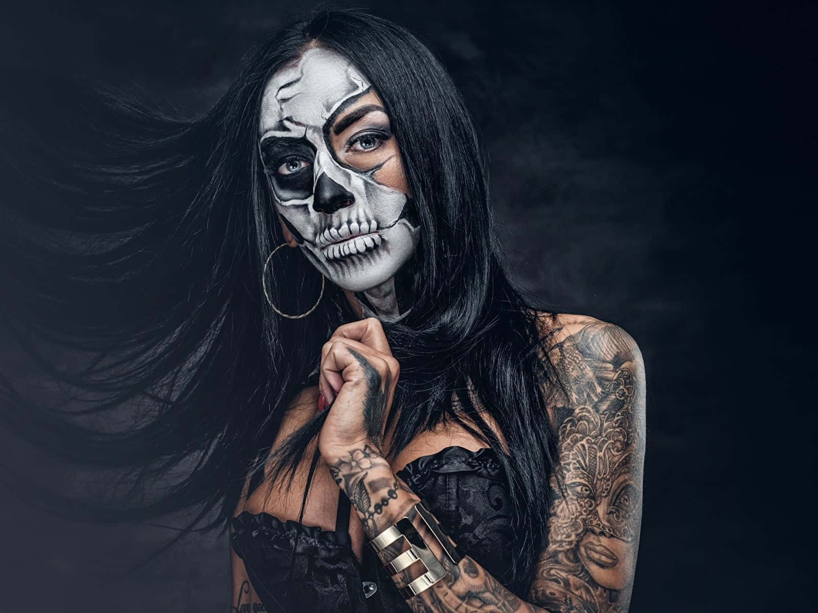 Day Of The Dead 桌布 壁紙 — 一位化著半骷髏妝容、帶有刺青的女性，在黑暗煙霧背景前擺姿勢，黑色長髮飄逸。