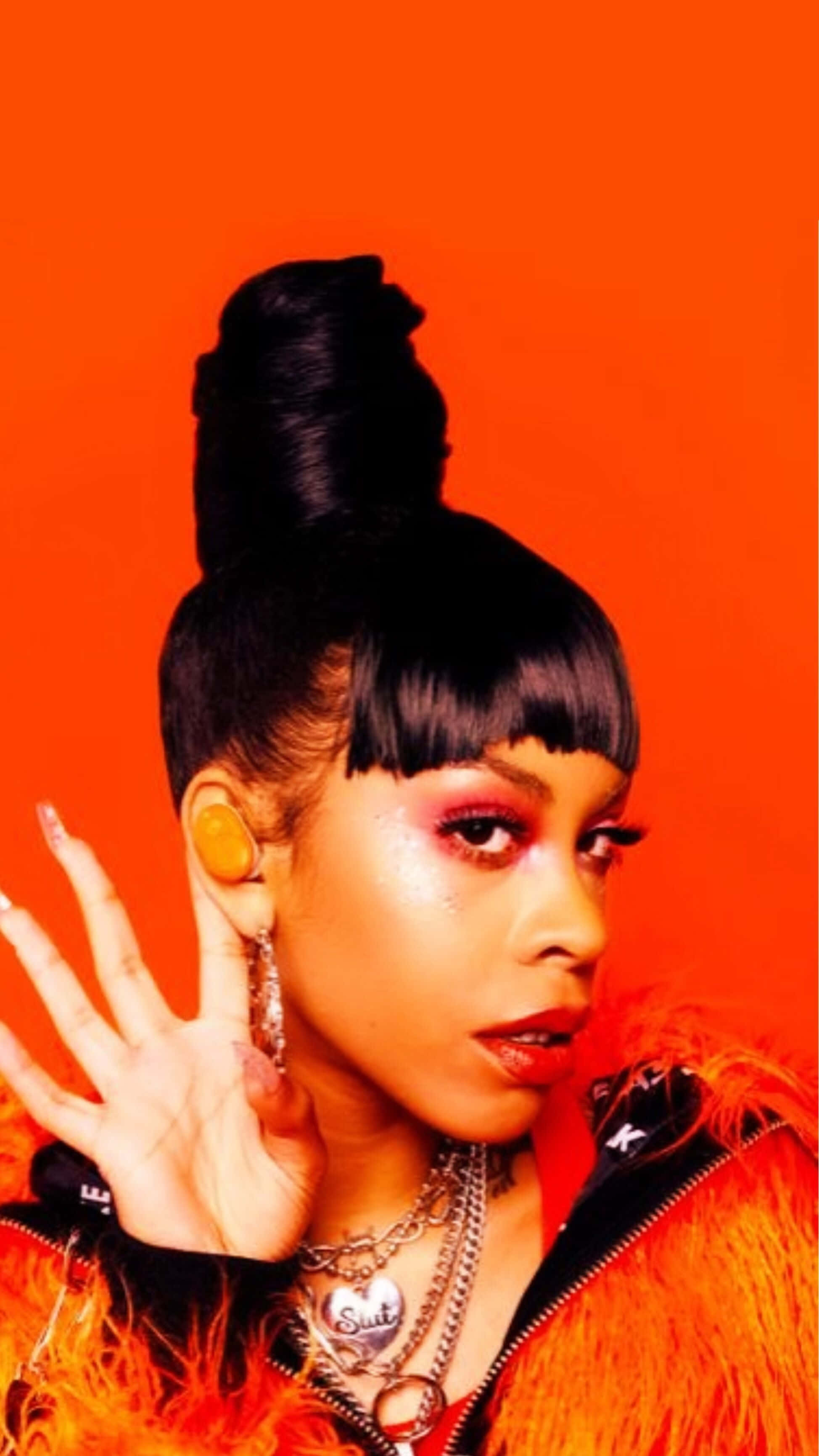 Rico Nasty 桌布 壁紙 — 一位化著大膽妝容、紮著高馬尾的女性，在鮮豔的橙色背景前穿著羽毛外套擺姿勢。
