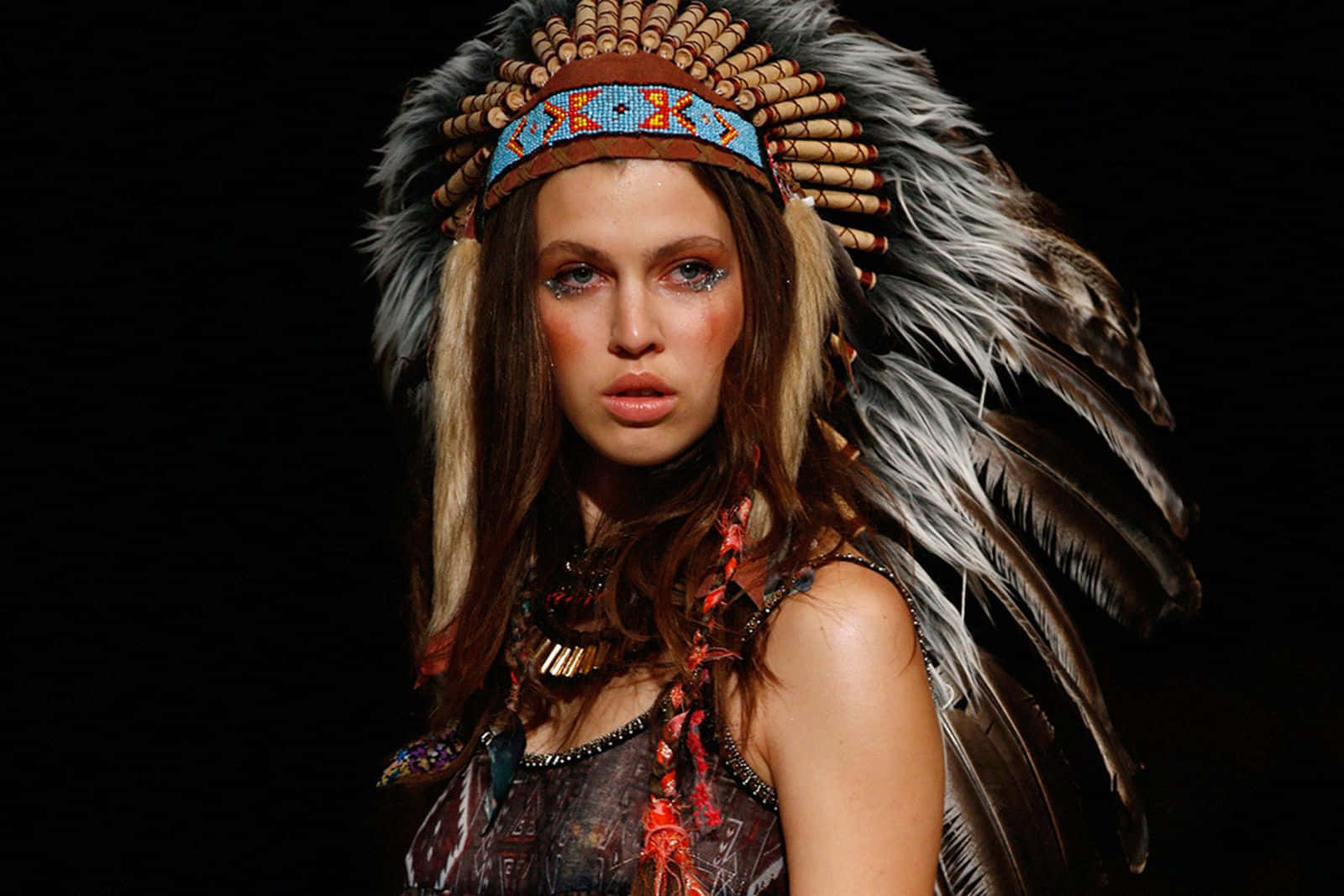 Native American 桌布 壁紙 — 一位化著搶眼妝容的模特兒，戴著精緻的羽毛頭飾和珠飾配件，以黑色背景襯托。