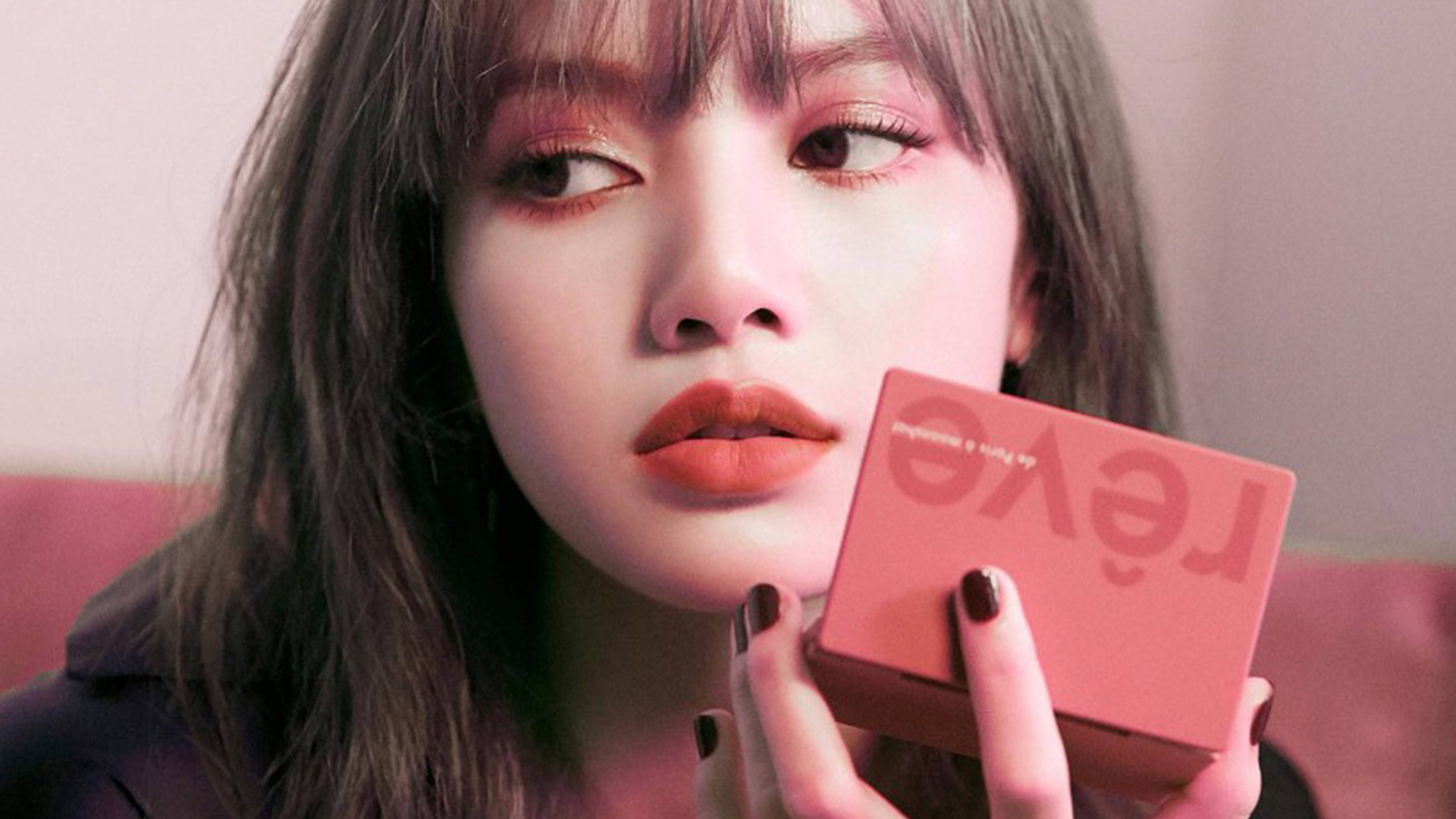 Lalisa 桌布 壁紙 — 一位化著柔和粉紅妝容、深色指甲的女性，在柔和的粉紅背景前拿著珊瑚色腮紅粉盒。