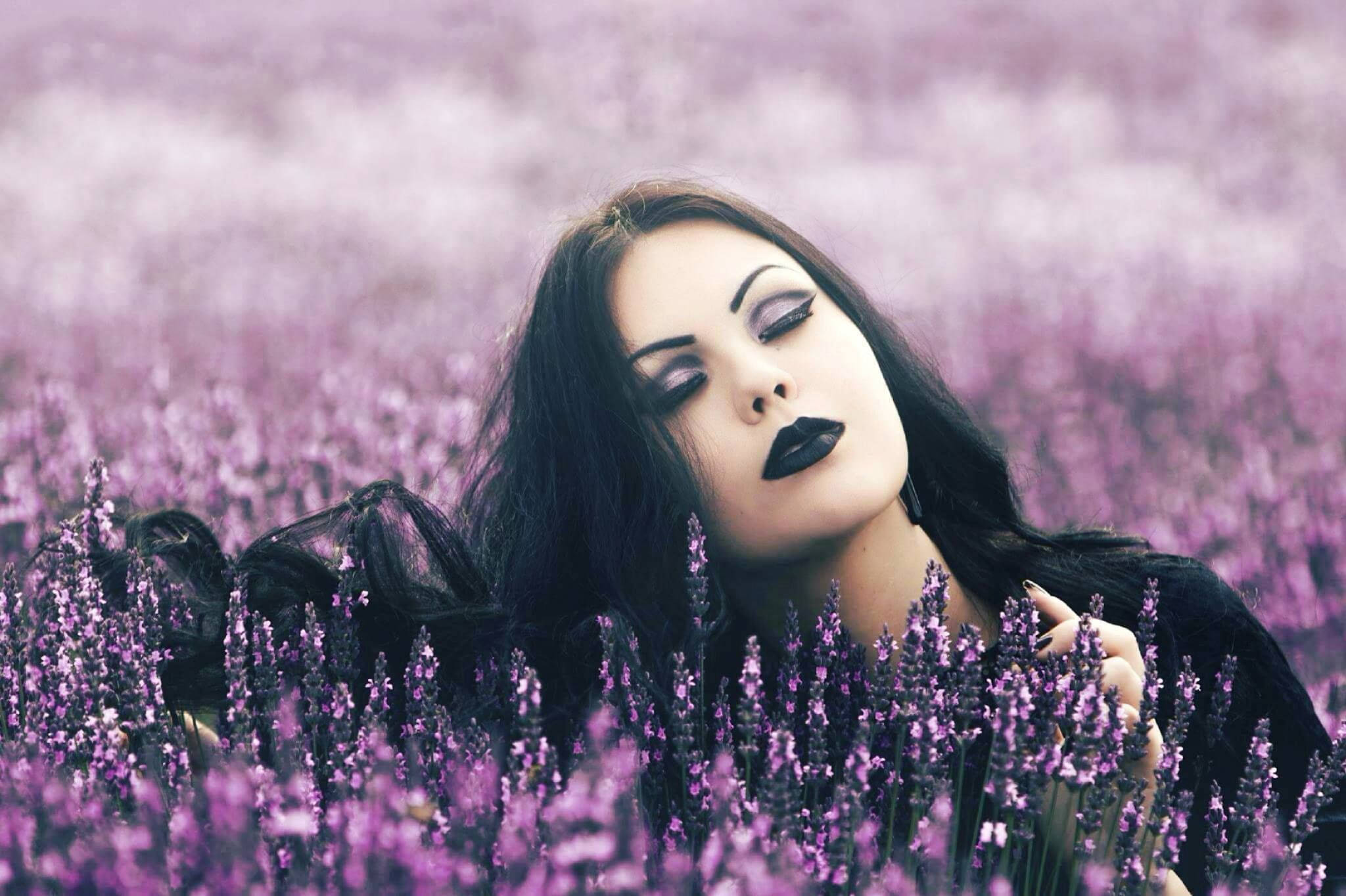 Goth 桌布 壁紙 — 一位化著深色妝容、深色頭髮的女性躺在柔和焦點的田野中，周圍是鮮豔的紫色薰衣草花。