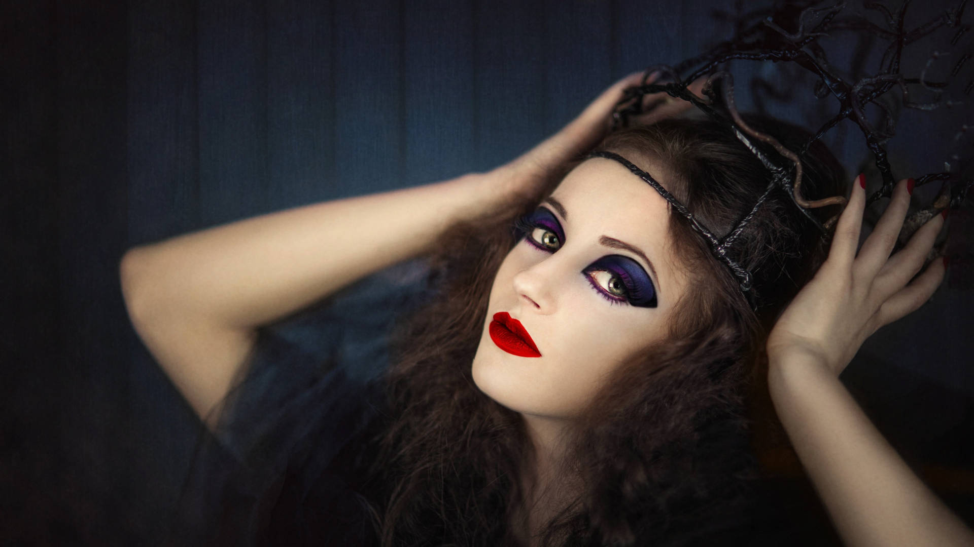 Witchy 桌布 壁紙 — 一位化著深色妝容和紅唇的女性，雙手放在頭髮上，在陰影藍色背景前擺姿勢。