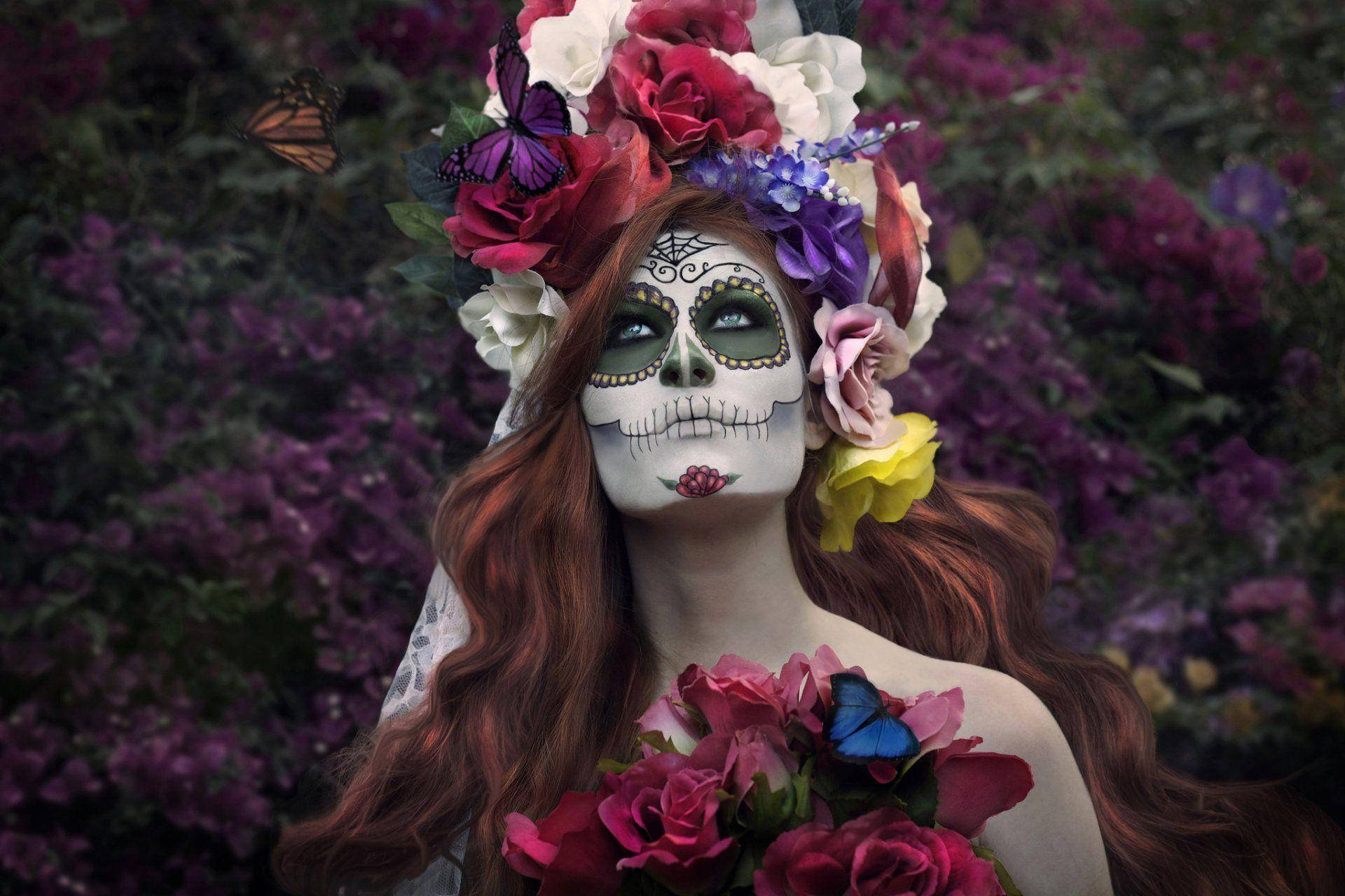 Dia De Los Muertos 桌布 壁紙 — 一位化著糖骷髏妝容、戴著花冠的女性，站在紫色花朵中，蝴蝶在她周圍翩翩起舞。
