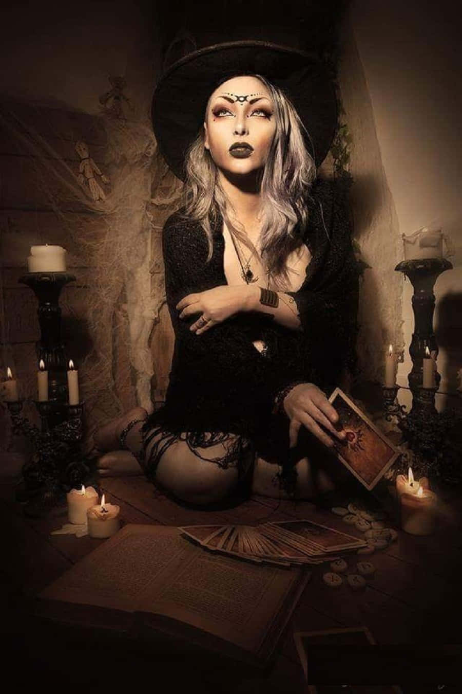 Witch Costumes 桌布 壁紙 — 一位化著蒼白妝容、戴著黑色帽子的女巫，在燭光昏暗的環境中跪著，周圍散落著塔羅牌和一本打開的魔法書。