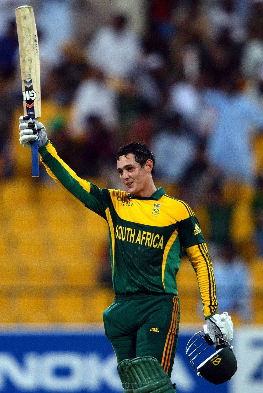 quinton de kock 桌布 壁紙 — 一位南非板球運動員在充滿歡呼球迷的體育館中，微笑著高舉球棒，慶祝勝利。