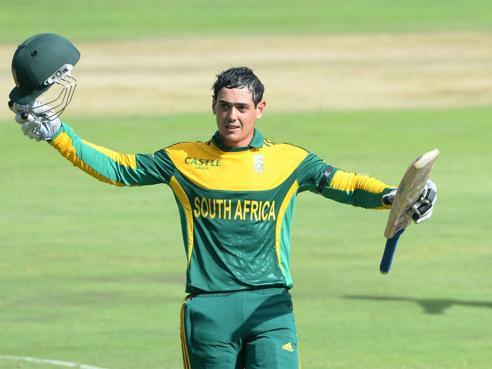 quinton de kock 桌布 壁紙 — 一位南非板球運動員在陽光明媚的綠色球場上，高舉頭盔和球棒，勝利慶祝。