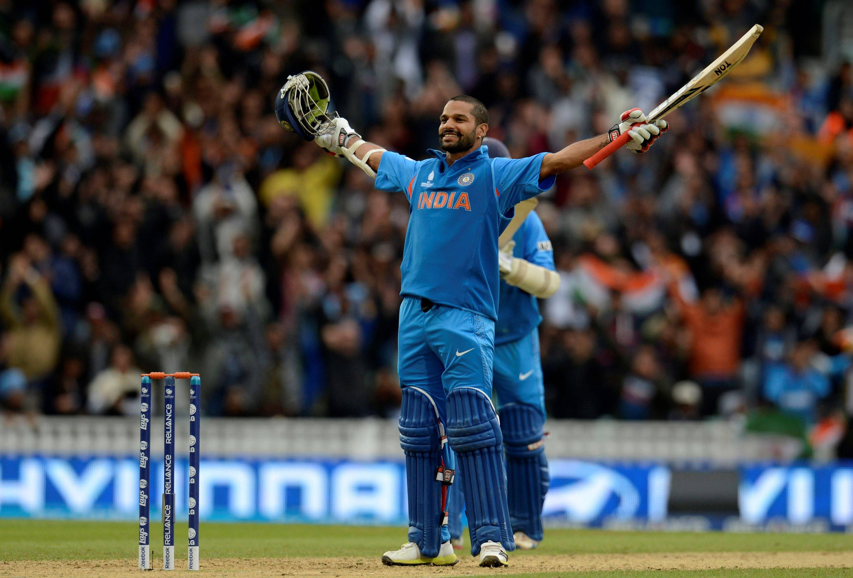 shikhar dhawan 桌布 壁紙 — 一位印度板球運動員在陽光明媚的球場上勝利慶祝，高舉球棒和頭盔，面對歡呼的球迷。